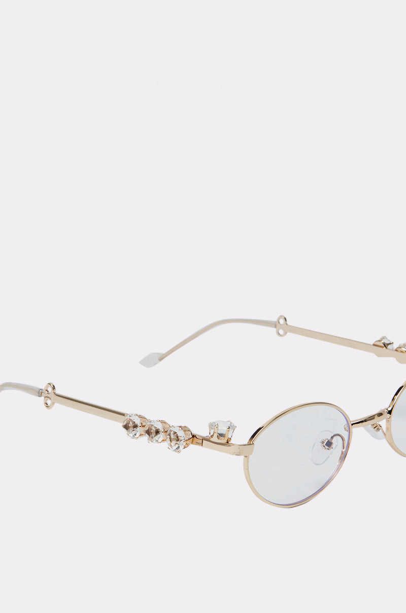 The PIERCED VIBES CLEAR GLASSES feature a gold-tone metal oval frame and clear lenses, with decorative crystal accents along the arms, showcased on a light grey background. 4