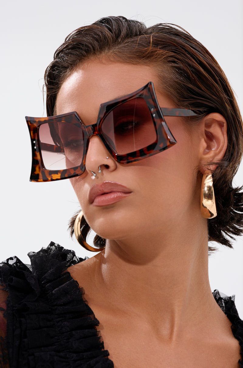 A woman in oversized SHELLY SUNGLASSES with retro tortoiseshell frames, gold hoops, and a black textured top. She has slicked-back short hair, a septum piercing, and gazes to the side against a light background. 1