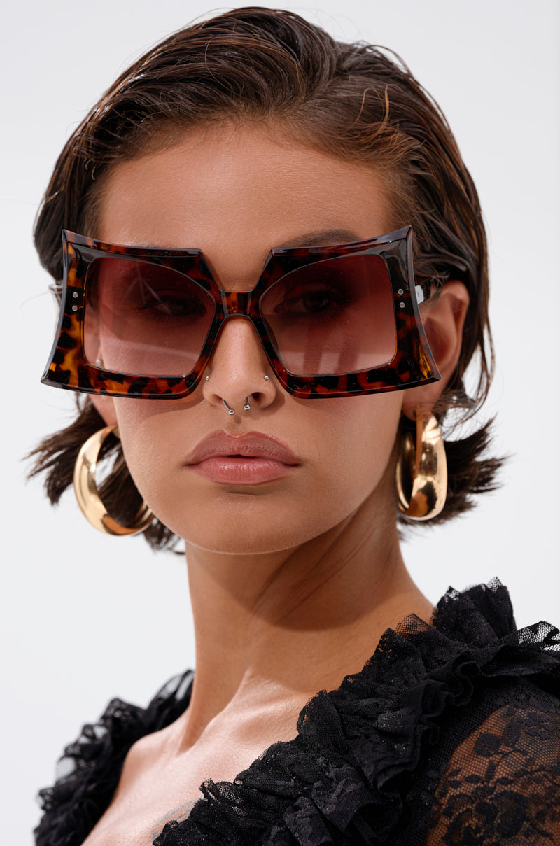 A person with short brown hair wears oversized, square tortoiseshell SHELLY SUNGLASSES, large gold hoop earrings, and a black lace top. They have a neutral expression and a septum piercing against a plain light gray background.
