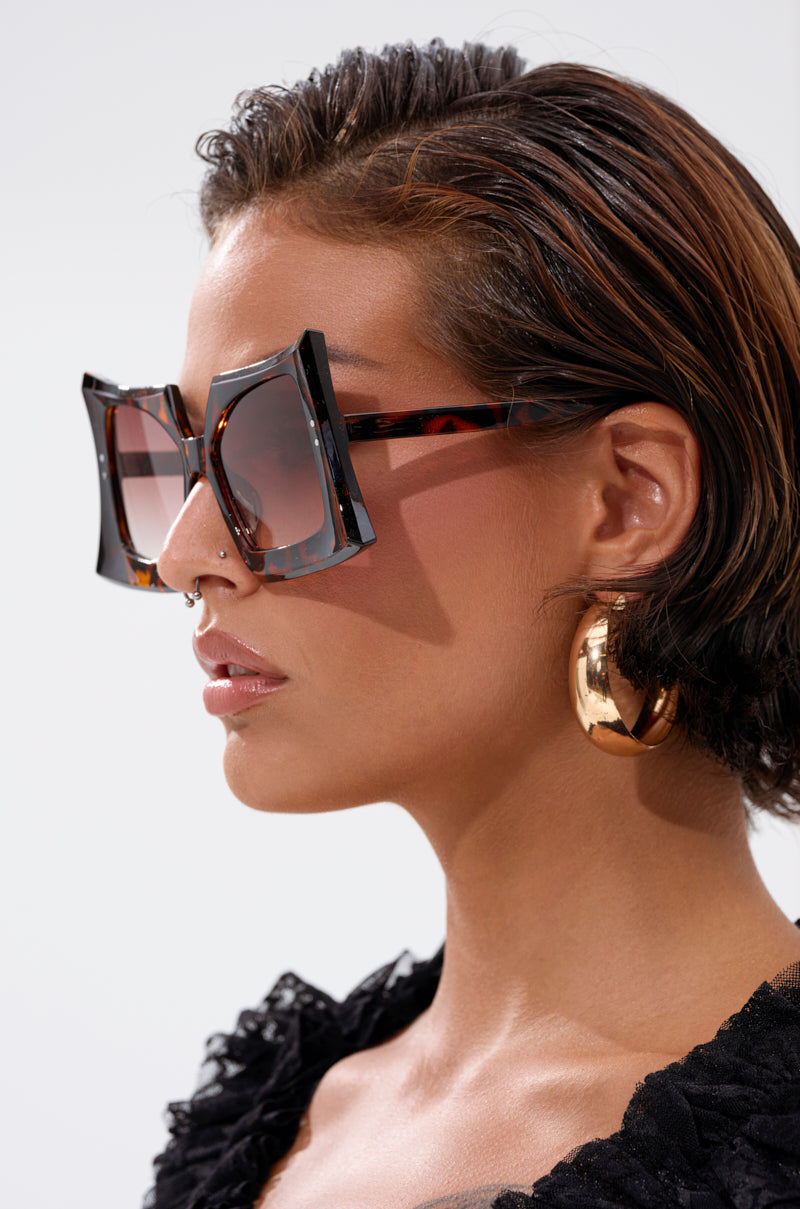 A person with short, slicked-back hair wears SHELLY SUNGLASSES—large, square frames—paired with gold hoop earrings and a textured black top, shown in profile against a plain light background.