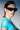 A woman with slicked-back hair models HERE FOR THE VIBES SUNGLASSES, paired with a sheer turquoise top and silver chain necklace, posing against a neutral backdrop under dramatic lighting.