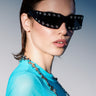 A woman with slicked-back hair models HERE FOR THE VIBES SUNGLASSES, paired with a sheer turquoise top and silver chain necklace, posing against a neutral backdrop under dramatic lighting.