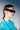 A person with slicked-back brown hair wears HERE FOR THE VIBES SUNGLASSES, a sheer turquoise top, and a beaded necklace, posing against a gradient light gray background.