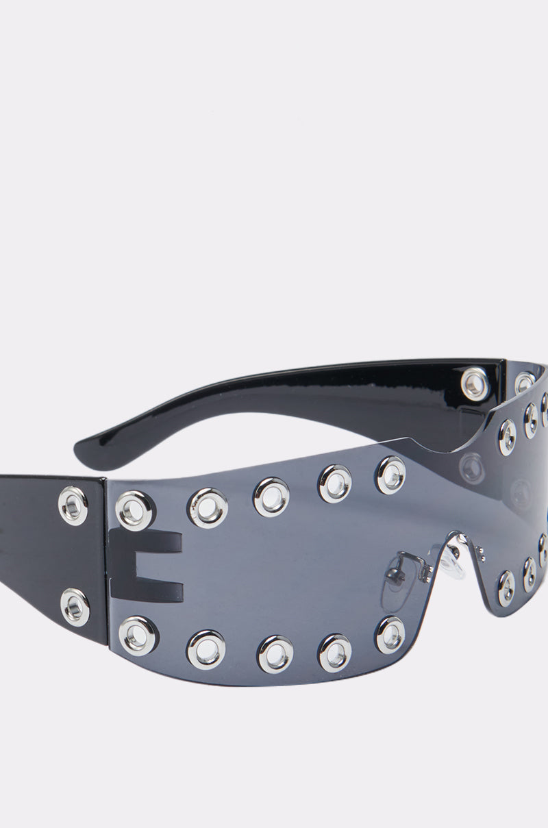 The HERE FOR THE VIBES SUNGLASSES feature a black futuristic wraparound frame, a single curved tinted visor lens, and silver grommets along the top and bottom edges for a bold, fashion-forward look. 5
