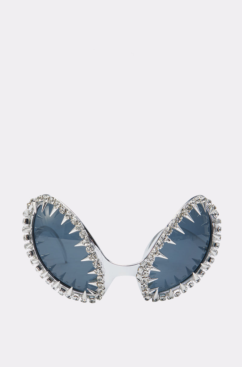 BUGGIN BLING SUNGLASSES feature blue-tinted, wing-shaped lenses outlined in rhinestones with a jagged silver edge, creating a bold open-jaw effect. These avant-garde sunglasses stand out against a white background.