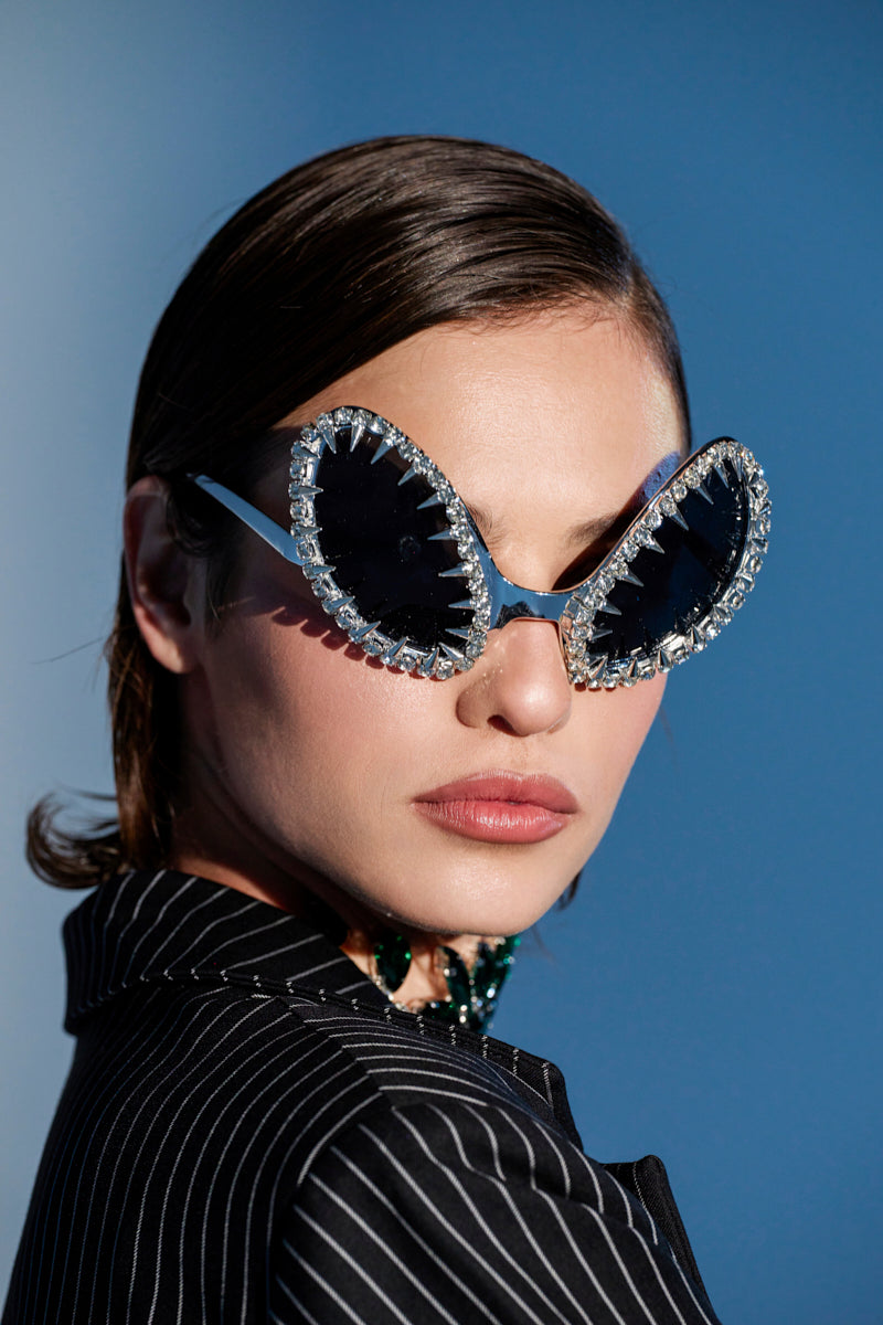 A woman with short brown hair confidently poses against a blue background wearing BUGGIN BLING SUNGLASSES and a black pinstripe blazer.
