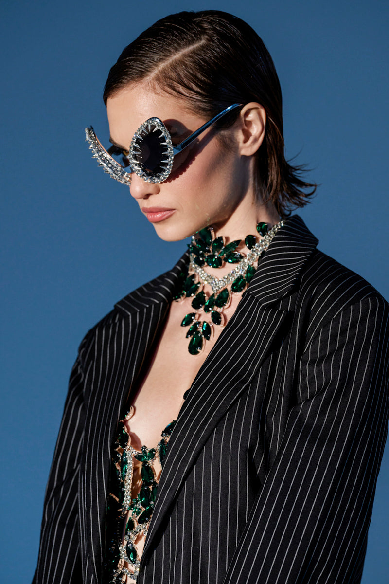 A person with slicked-back hair wears BUGGIN BLING SUNGLASSES, a statement green gemstone necklace, and a pinstripe blazer against a blue background.