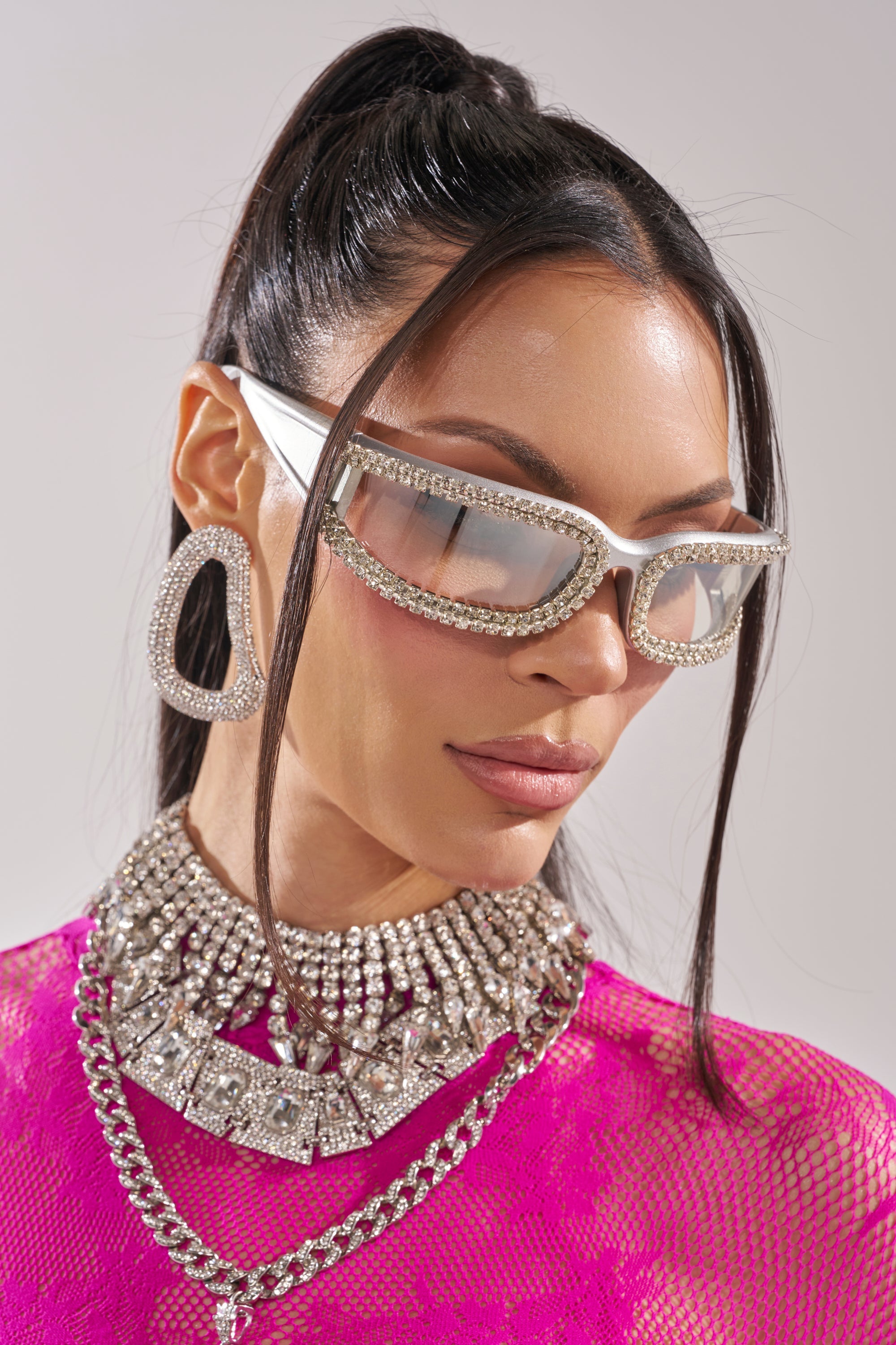 A woman with long dark hair in a high ponytail wears NIKKI SUNGLASSES, bold geometric earrings, layered rhinestone necklaces, and a bright pink lace top. 2