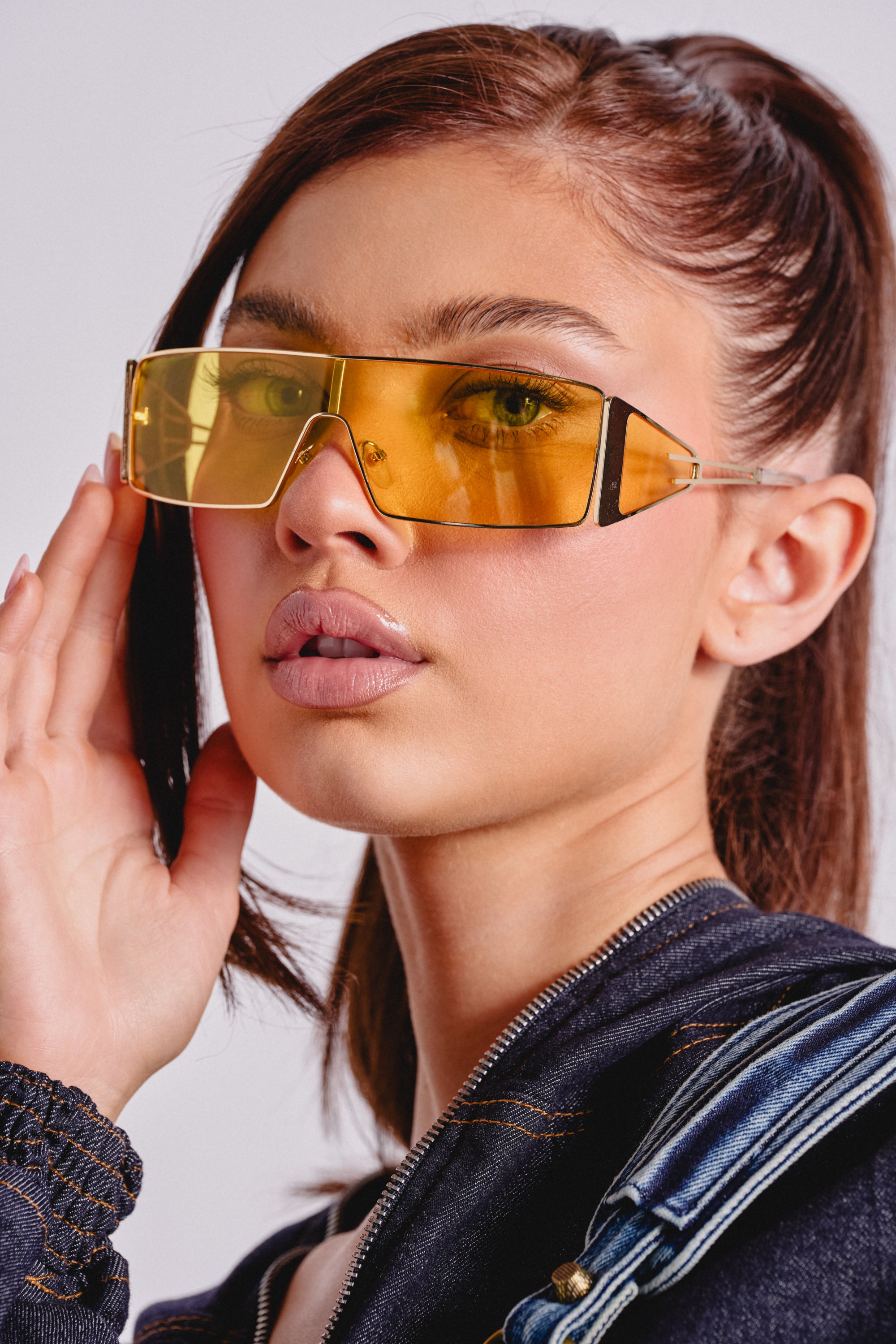 A woman with long brown hair in a ponytail wears LEMON TWIST SUNGLASSES—oversized yellow shield frames—and a dark zip-up jacket, looking into the camera while gently touching her statement eyewear. 0