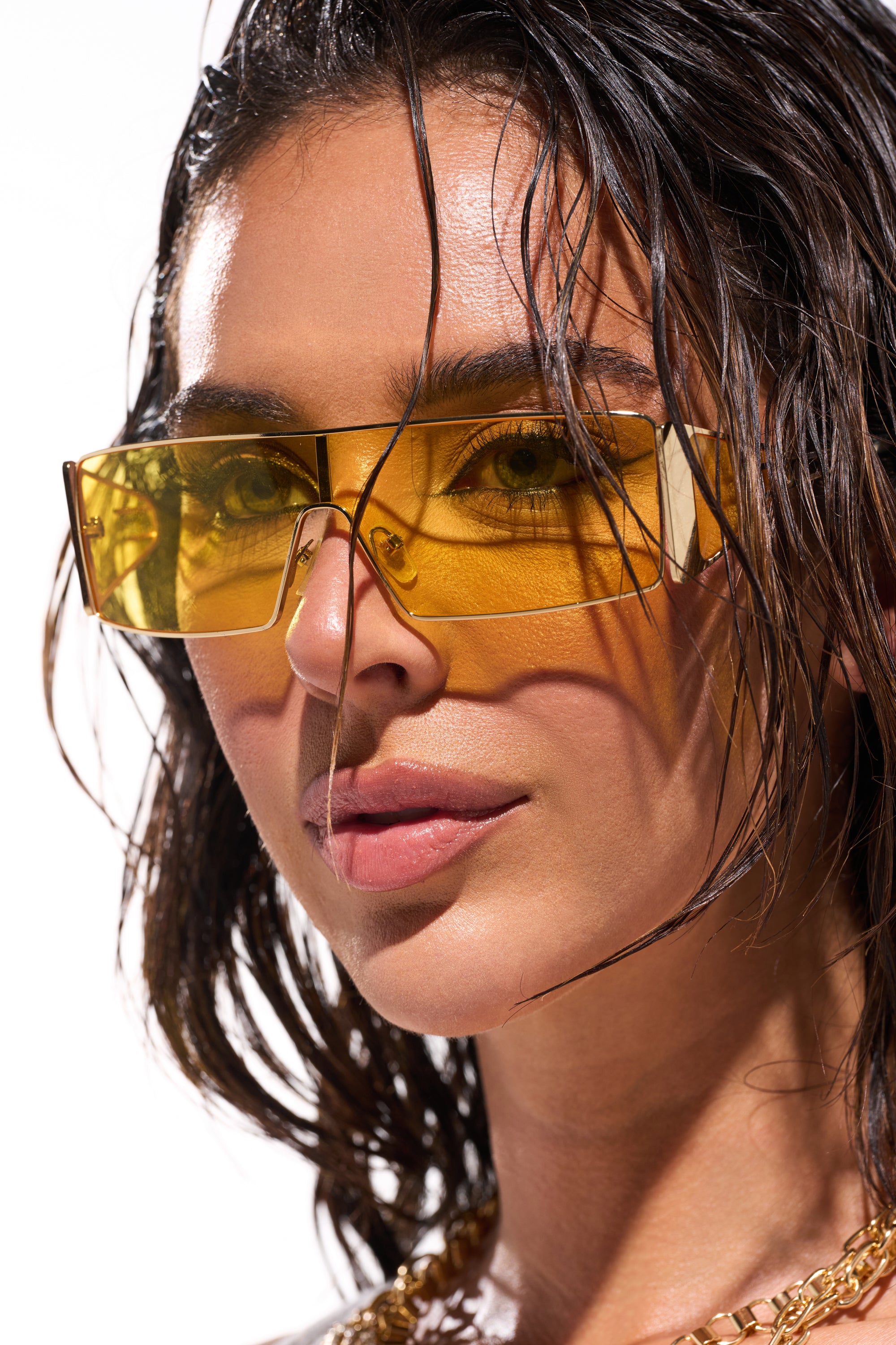 A close-up of a woman with wet, wavy dark hair wearing LEMON TWIST SUNGLASSES and a chunky gold chain necklace, as sunlight casts striking shadows on her face. 1