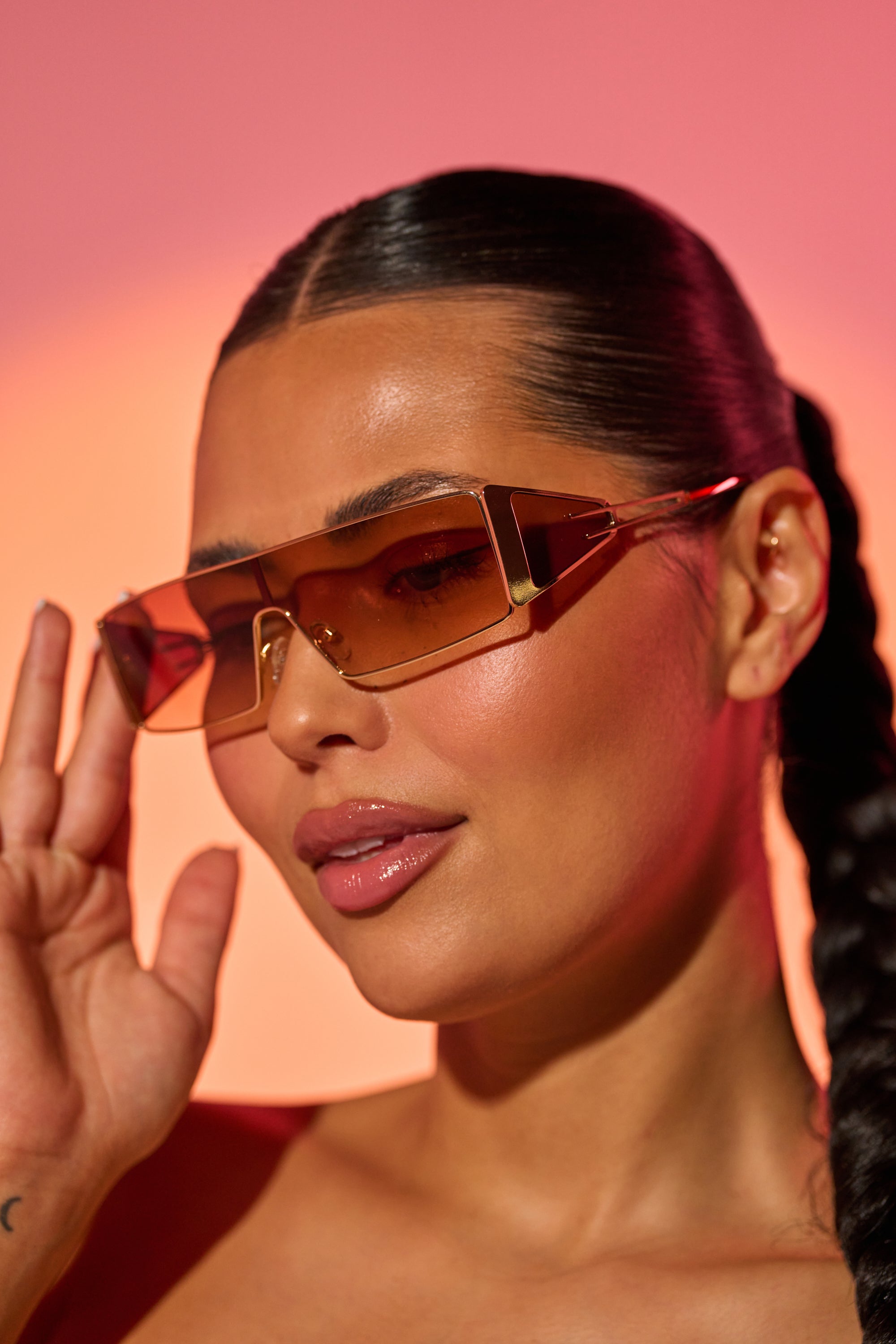 A woman with sleek dark hair in a braid wears VIXEN SUNGLASSES—retro shield shades with a gradient brown lens and gold-tone metal frame—and a glossy nude lip, posing against a warm pink-orange background. 1
