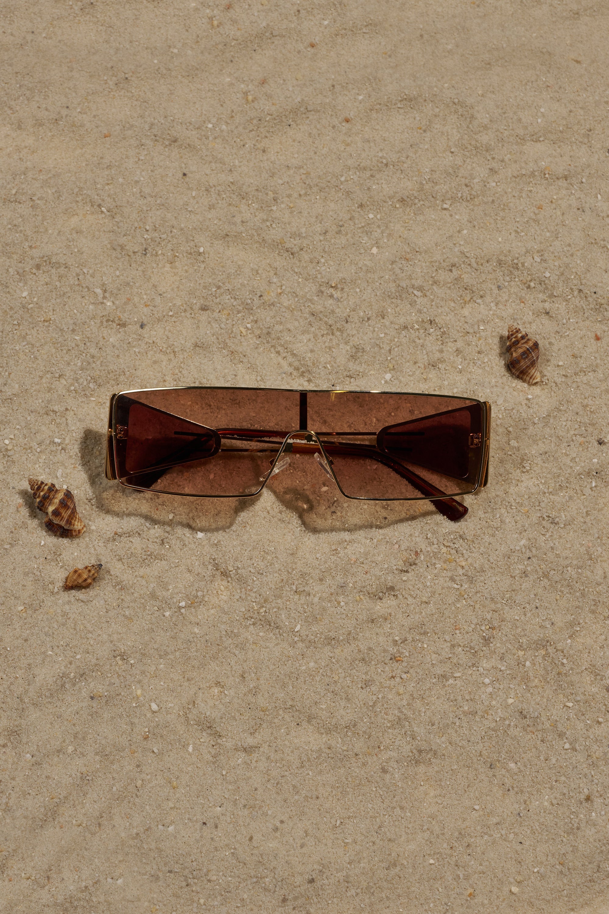 A pair of VIXEN SUNGLASSES with a gold-tone frame and gradient brown lenses rests on sandy ground, partially covered by sand, with four small seashells nearby. 0