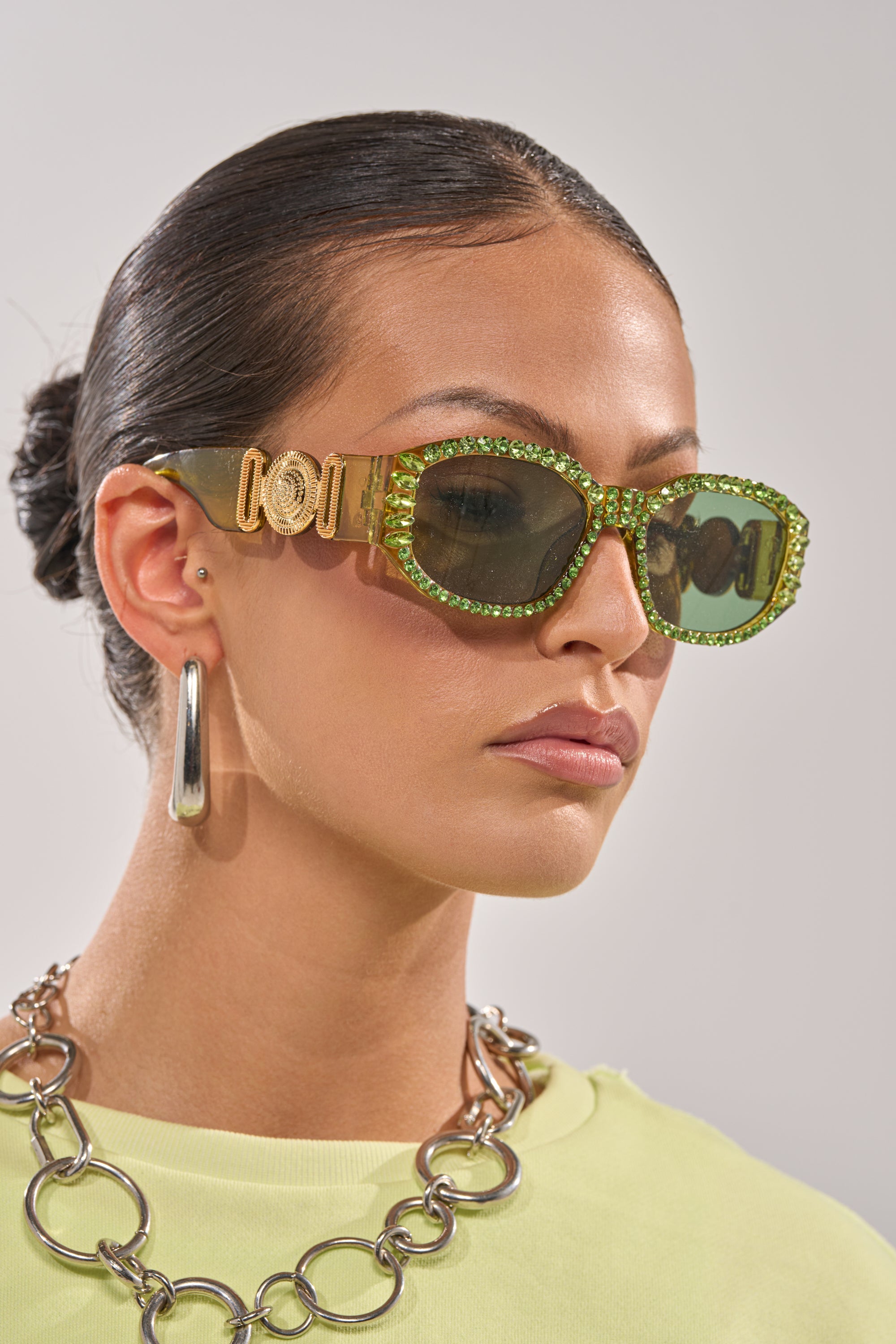 A woman with slicked-back hair wears CATCH MY VIBE SUNGLASSES, chunky silver hoop earrings, a large chain necklace, and a light yellow top, posing against a plain light background. 2