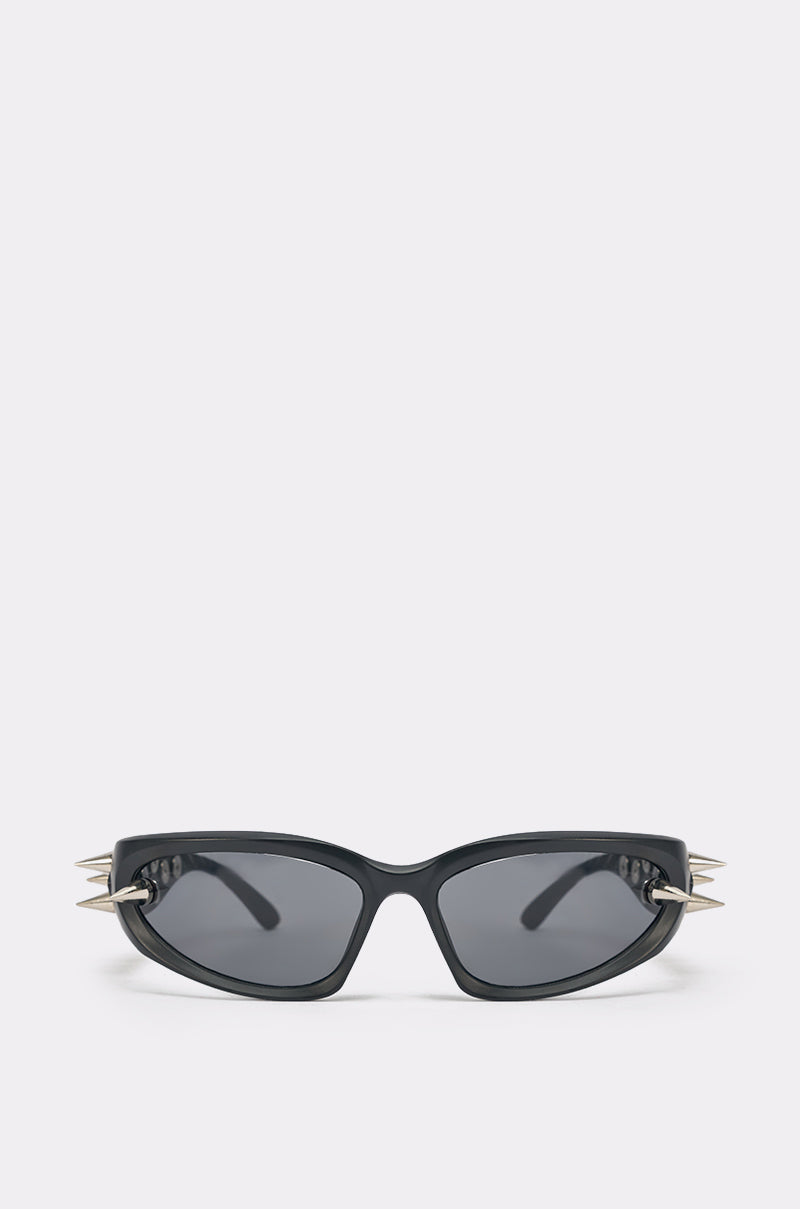 The PIERCE ME SUNGLASSES feature a bold black sculptural frame, dark tinted lenses, and striking metallic silver spikes on the sides, shown against a plain light background. 1
