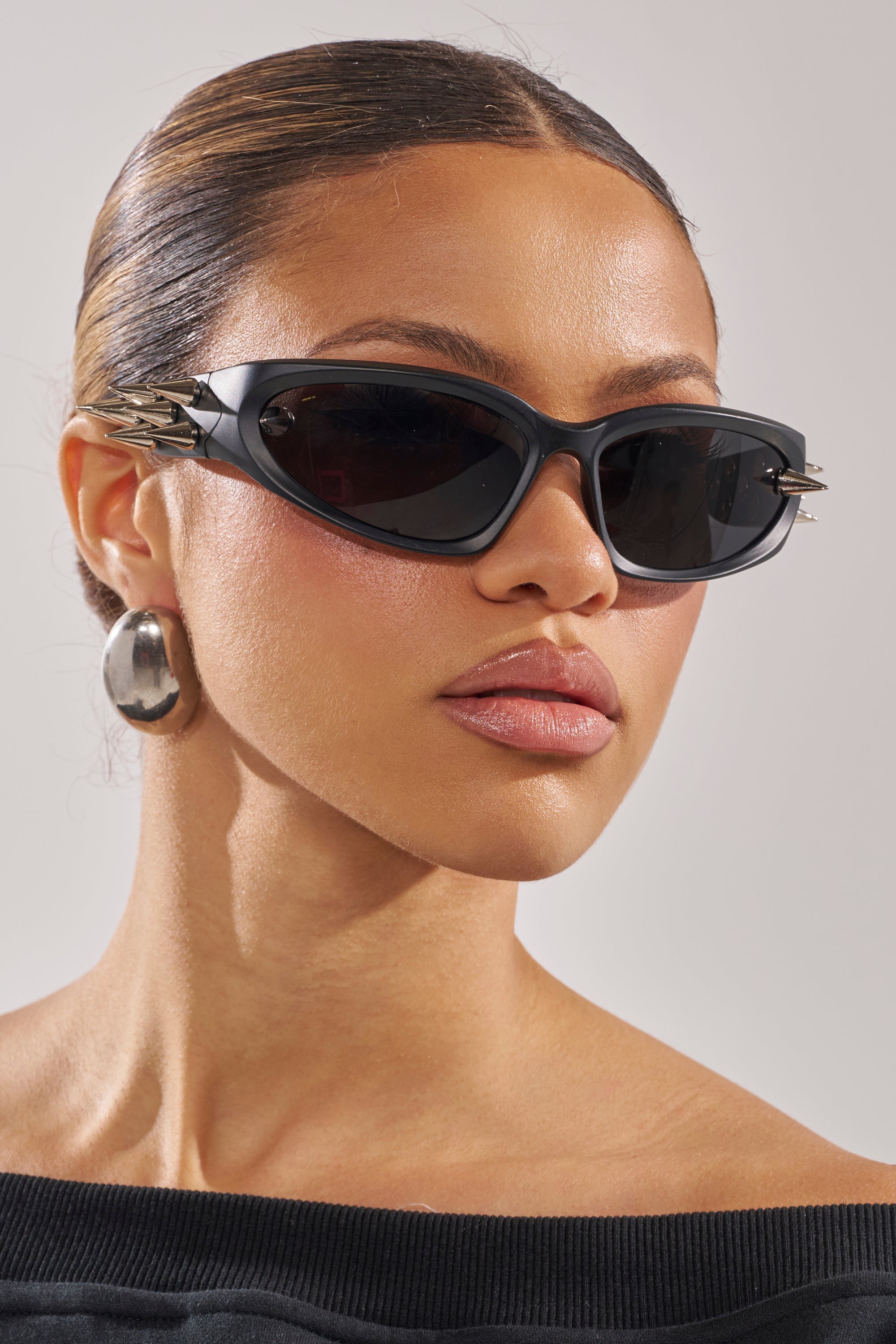 A woman with slicked-back hair wears PIERCE ME SUNGLASSES—black wrap frames with silver accents—plus large hoops and an off-the-shoulder black top, looking confidently ahead against a neutral background. 2