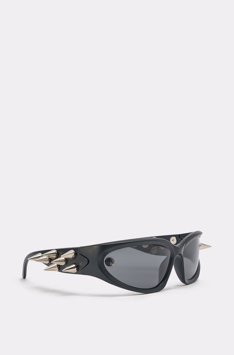 The PIERCE ME SUNGLASSES feature black statement frames with dark lenses and three silver spikes on each side, shown against a light grey background. 0