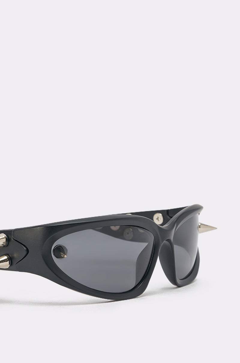 PIERCE ME SUNGLASSES feature a bold black sculptural wrap frame, dark oval lenses, and edgy silver stud hardware on the temples and corners, shown against a plain light background. 5