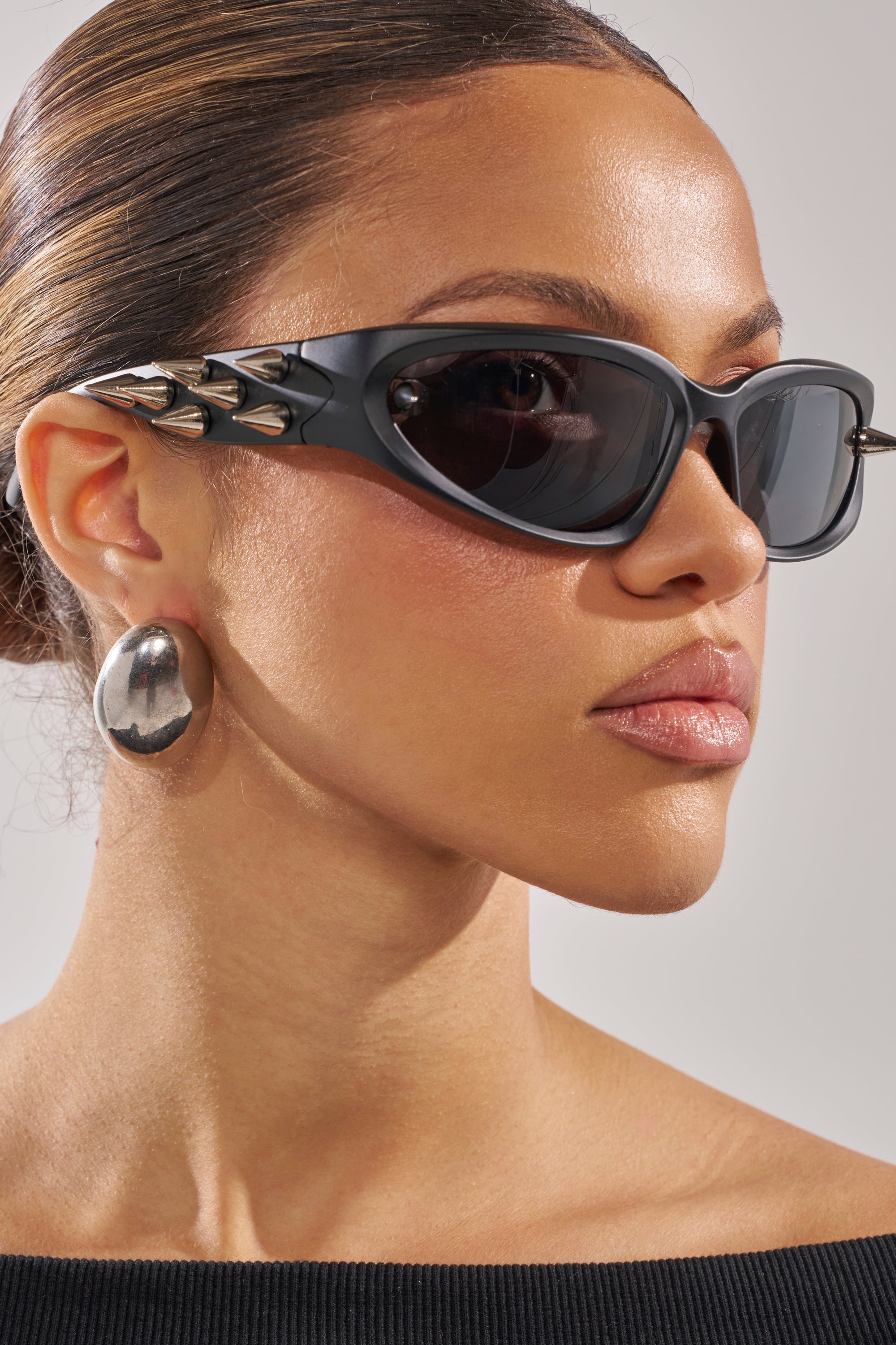 With her slicked-back hair and an off-the-shoulder black top, she exudes confidence in PIERCE ME SUNGLASSES—black wrap shades with metallic spikes—paired with large silver oval earrings. 4