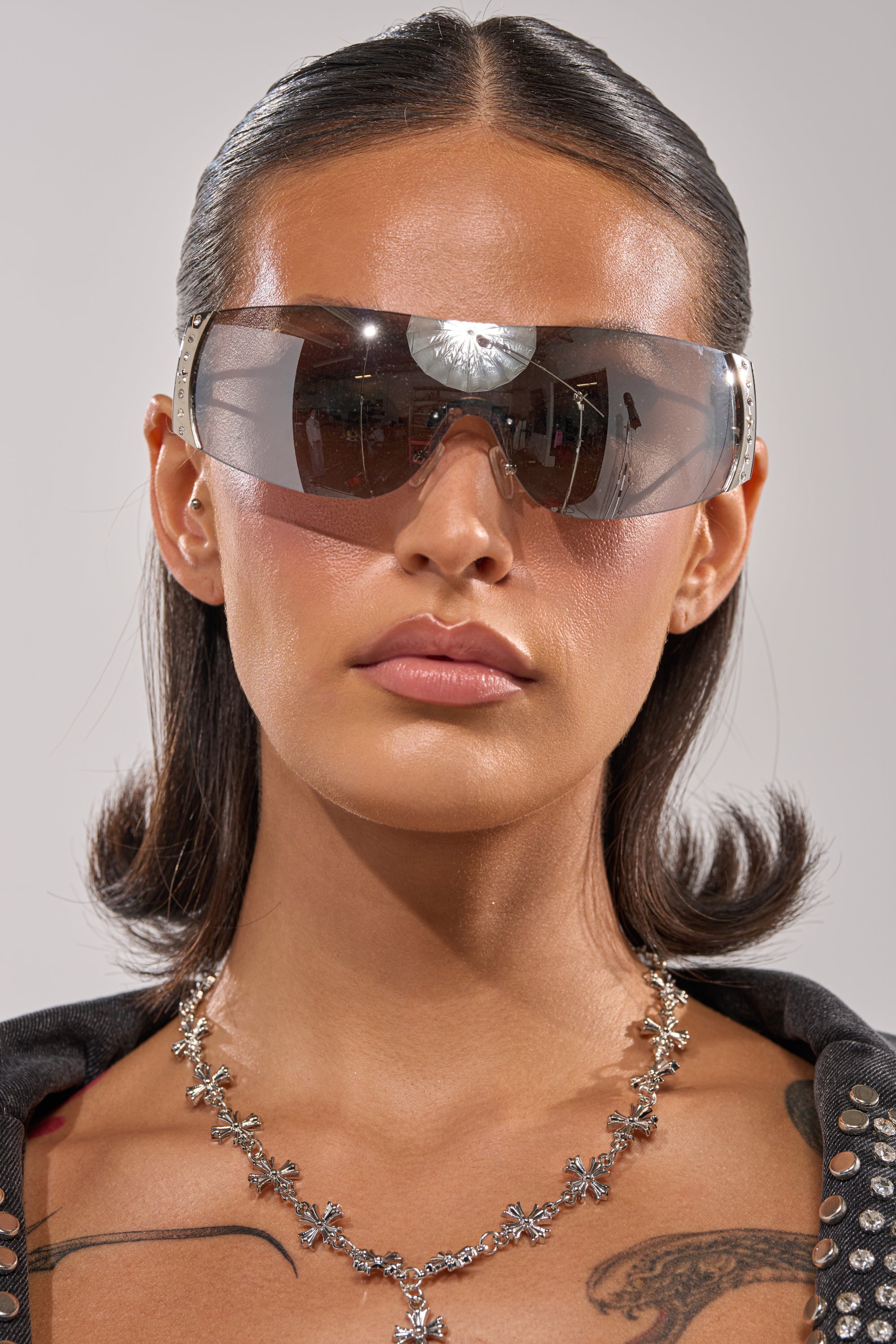 A woman with straight dark hair and visible shoulder tattoos poses against a plain background, wearing EVERY DAY SUNGLASSES—large shield frames with crystal accents—paired with a silver floral necklace and a black studded top. 4