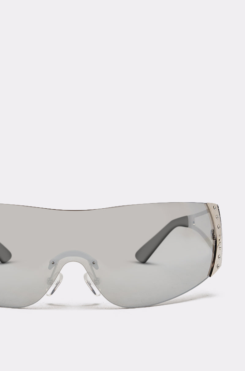 Close-up of EVERY DAY SUNGLASSES—oversized, frameless shield style with a single tinted lens and crystal-accented studs on the sides. Set against a plain, light background. 7