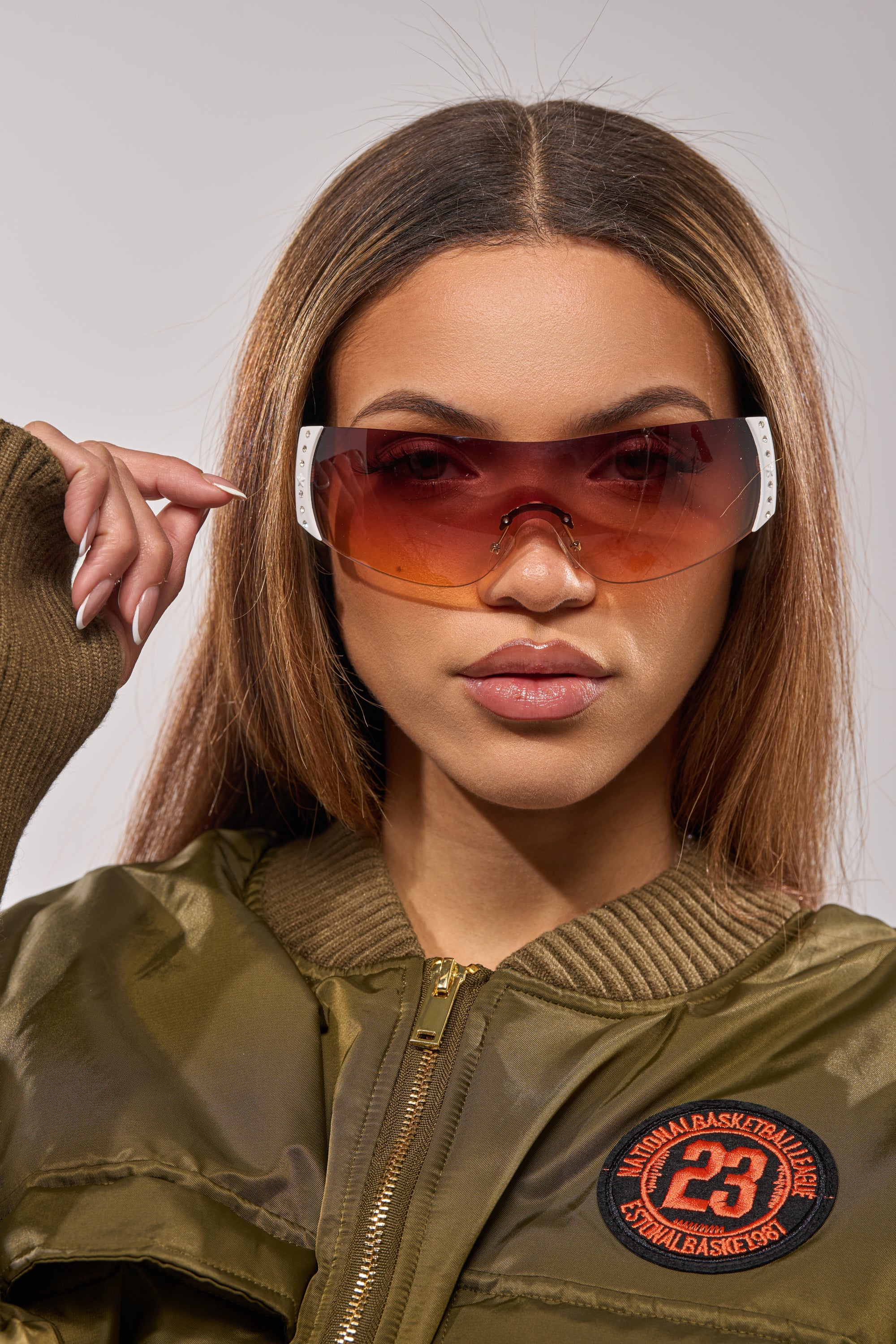 A woman poses confidently in NEUTRALEYES SUNGLASSES and an olive green bomber jacket with a basketball patch, straight hair framing her neutral expression as she holds one hand near her face. 4