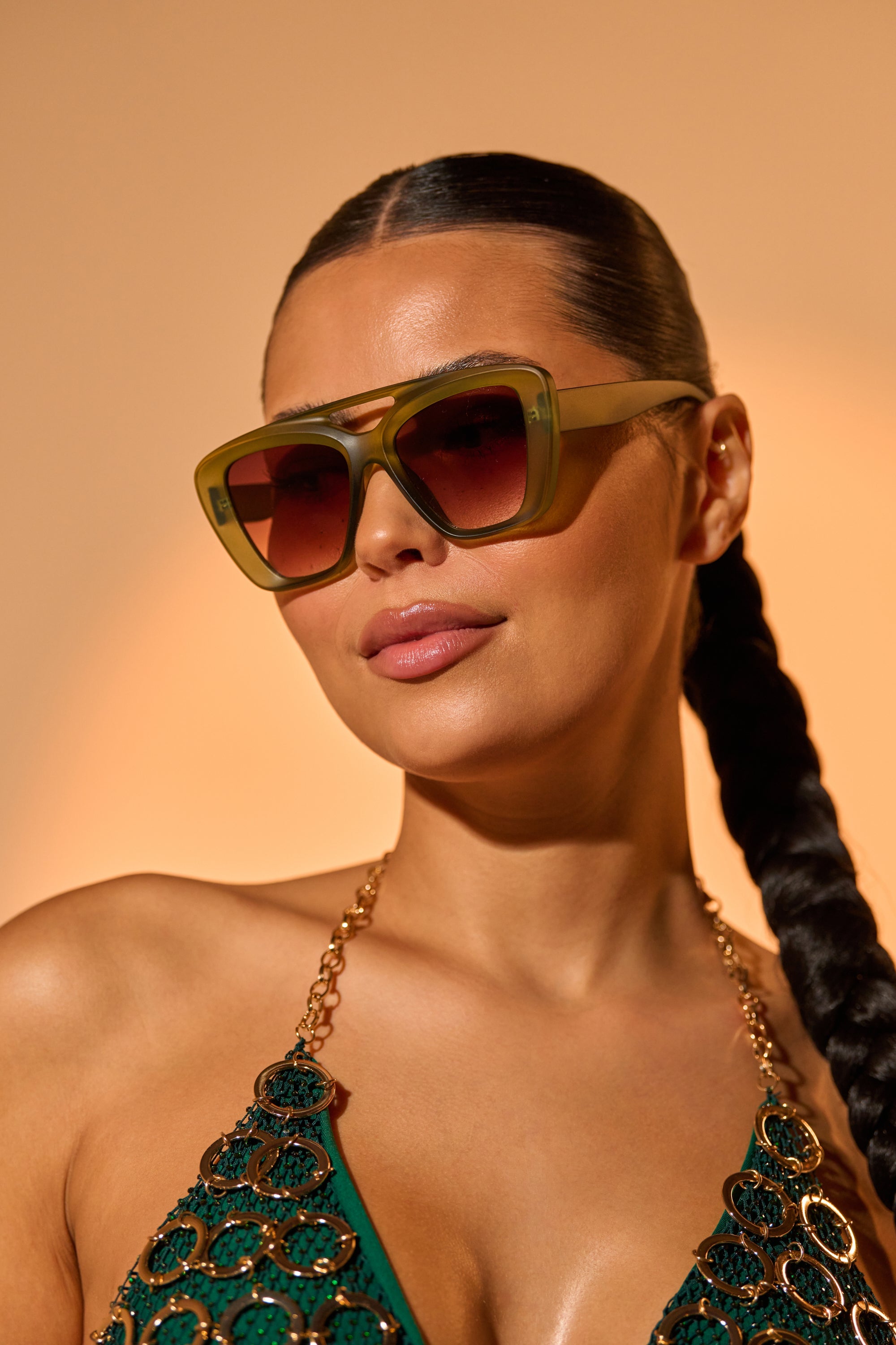 A woman with long dark hair in a sleek braid wears MAL SUNGLASSES—oversized olive-green frames—and a green top with gold chain straps, posing against a warm beige background. 0