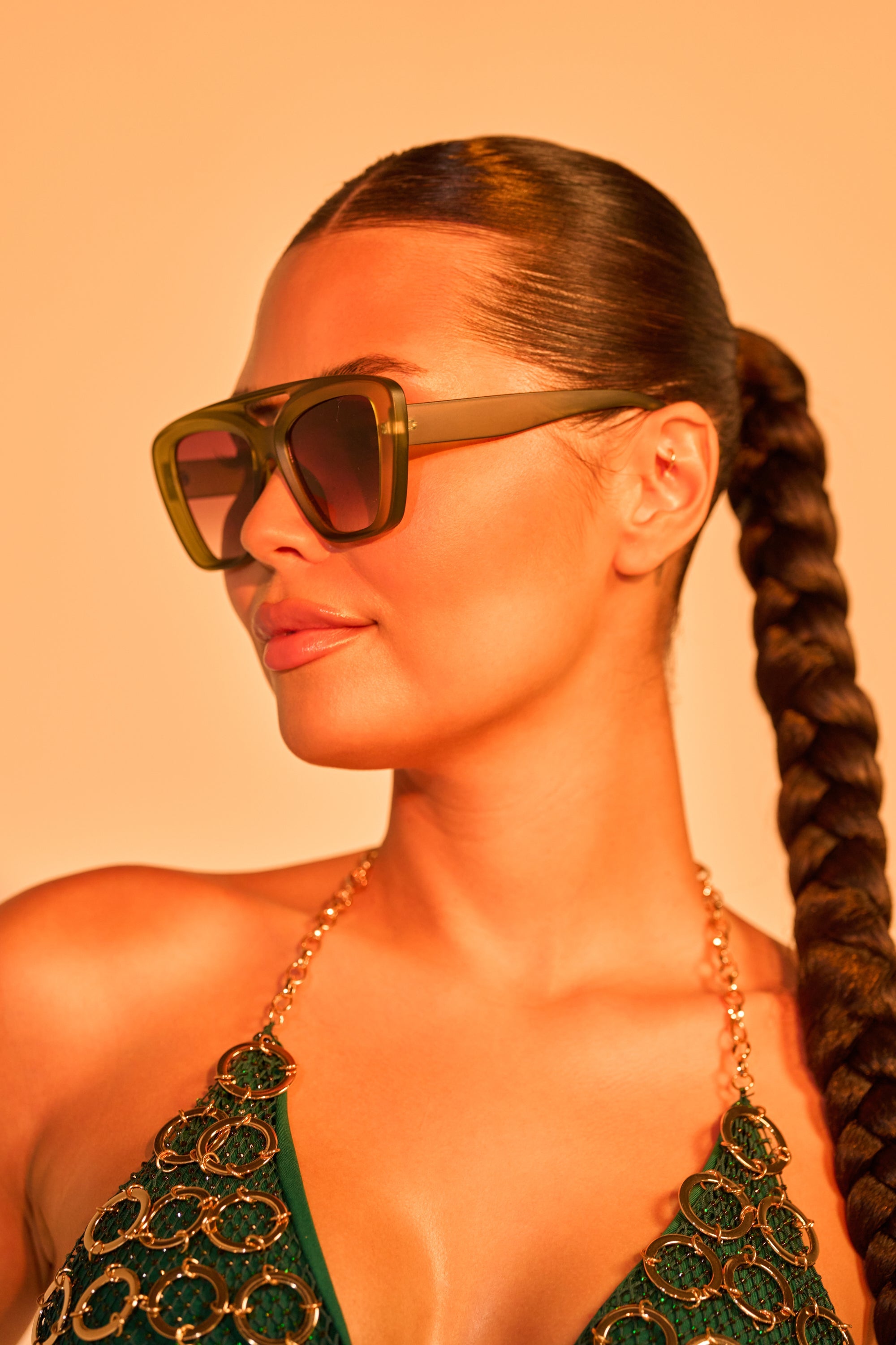 A woman with sleek, slicked-back hair in a long braid wears MAL SUNGLASSES and a green top with gold chain straps, set against a warm orange background. 1