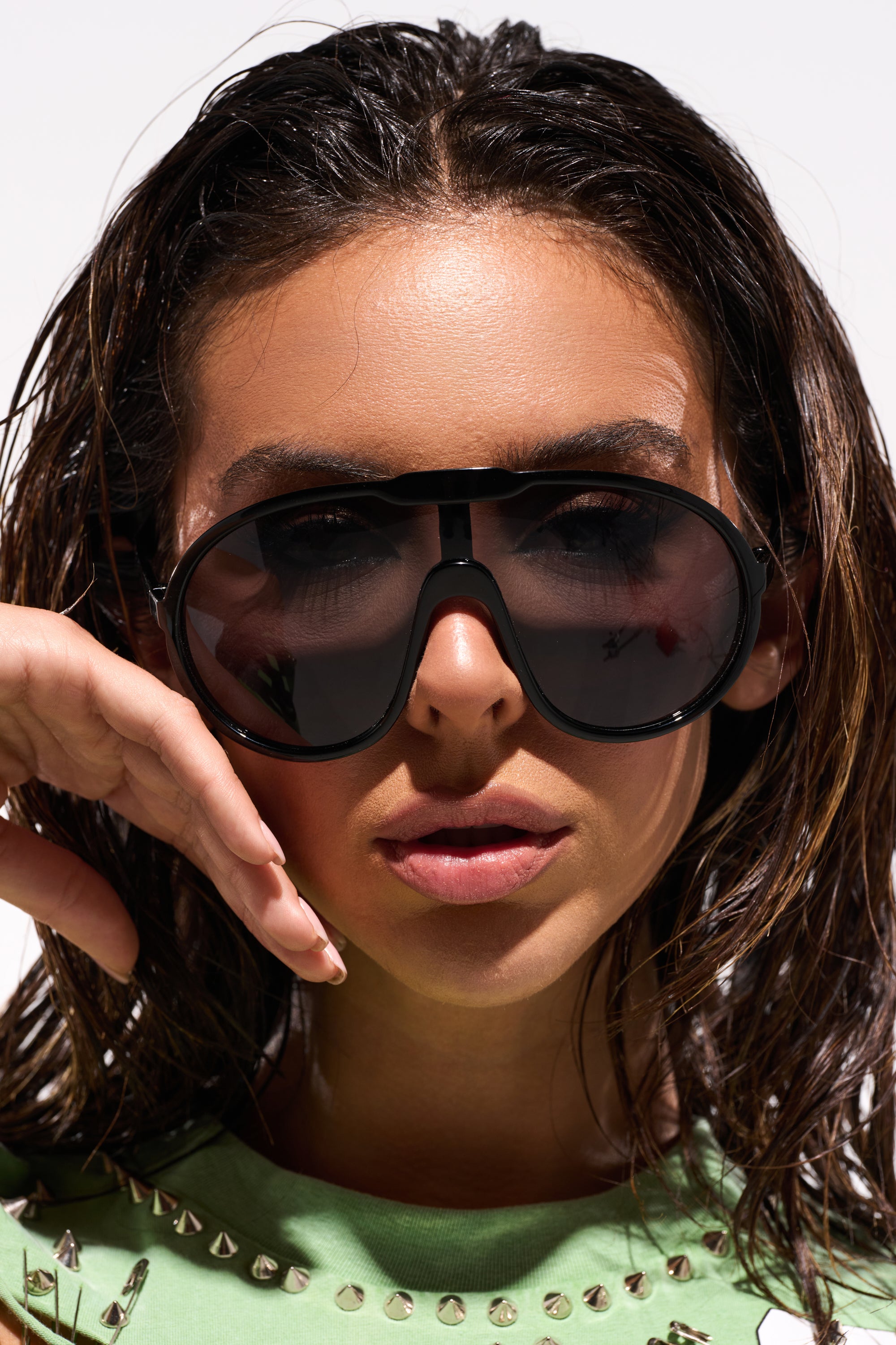 A woman with wet hair poses in BIGGER IS BETTER BLACK SUNGLASSES and a green top with silver studs, holding her hand near her cheek and looking at the camera against a plain background. 0