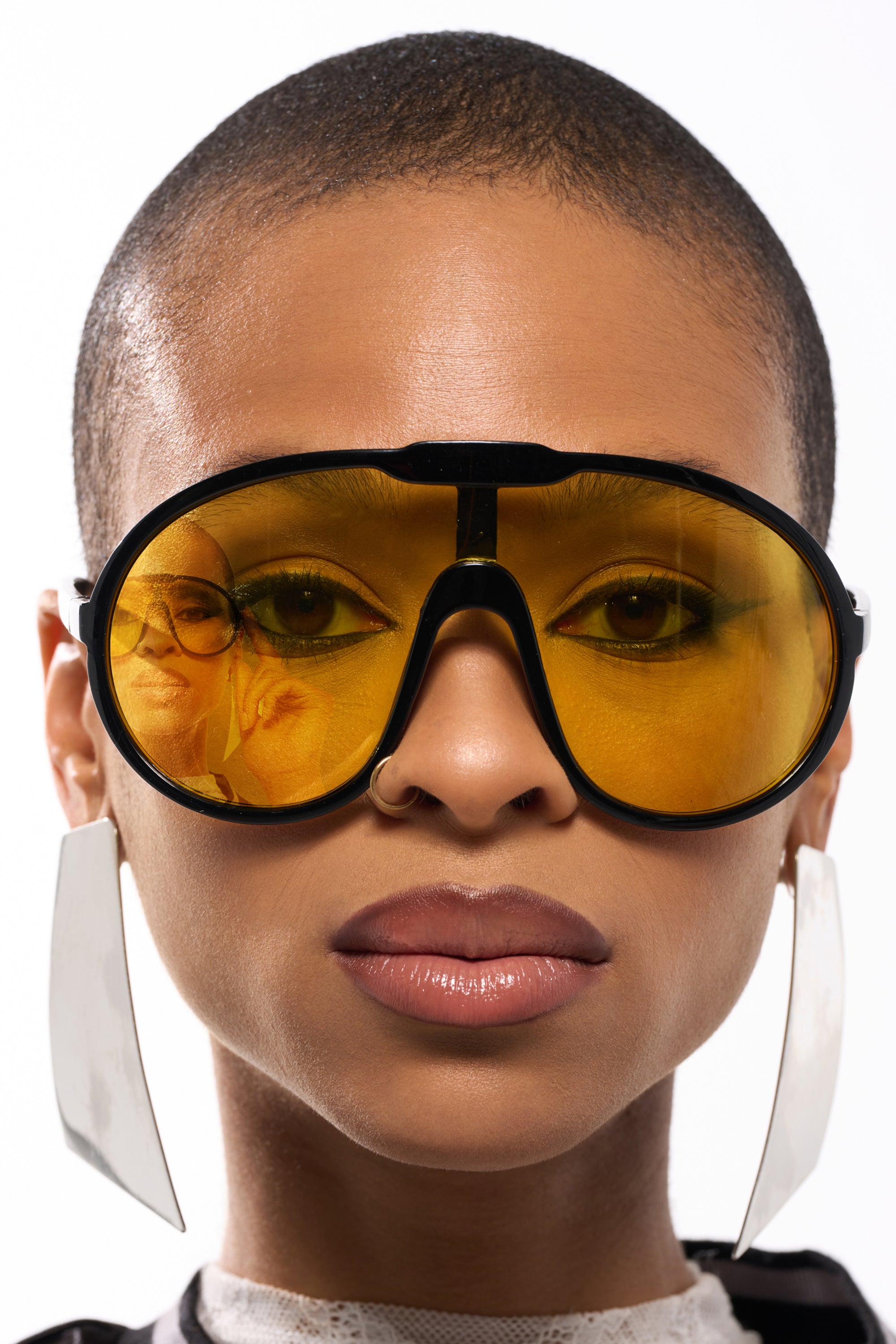 A person with a shaved head wears MOD SUNGLASSES featuring oversized wraparound yellow-tinted lenses and large rectangular earrings. Their neutral expression reflects two people in the retro-cool shades.