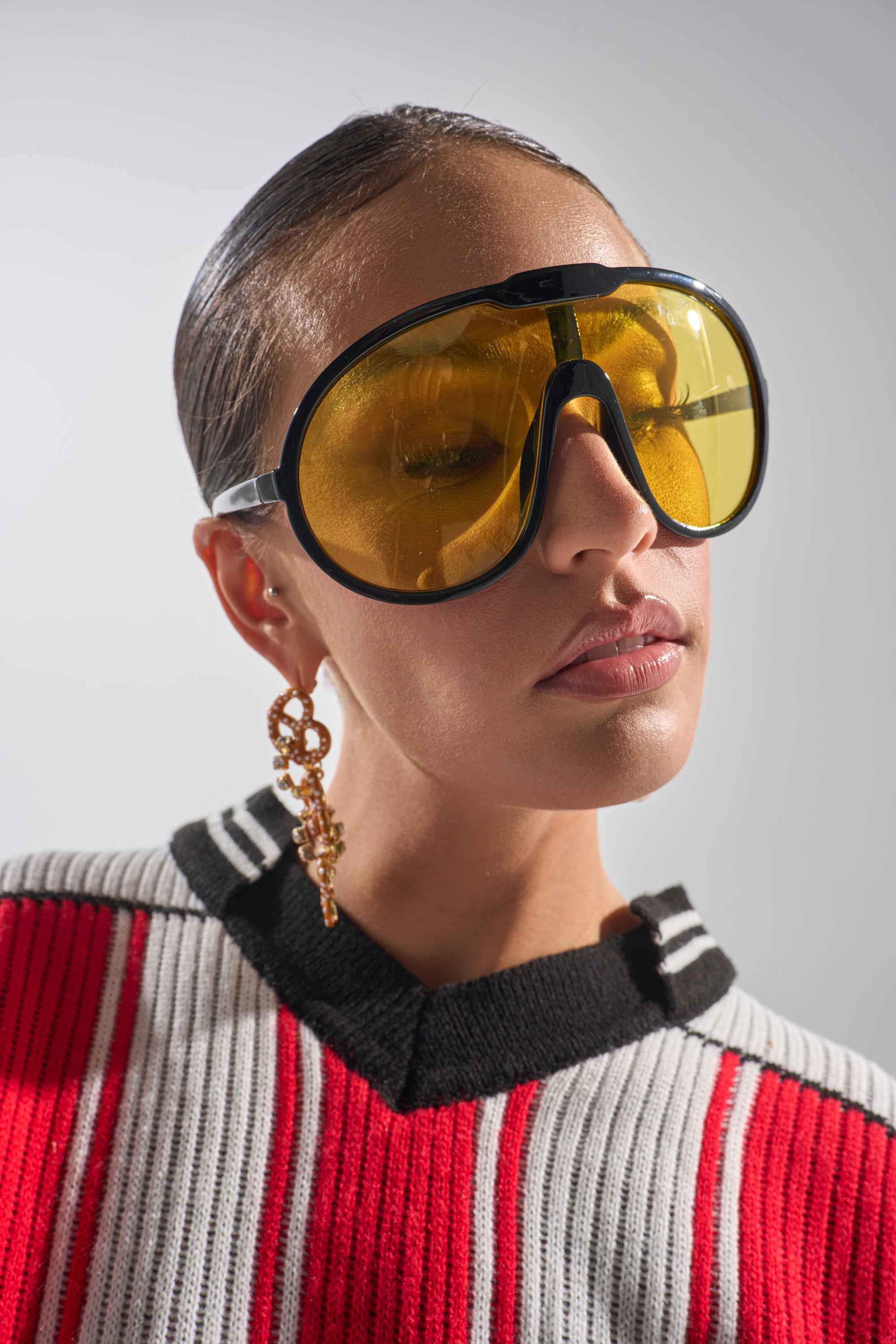 A person sports MOD SUNGLASSES—large, yellow-tinted retro shades—along with a chunky gold earring and a white sweater featuring red and black stripes, posing against a plain background. 0
