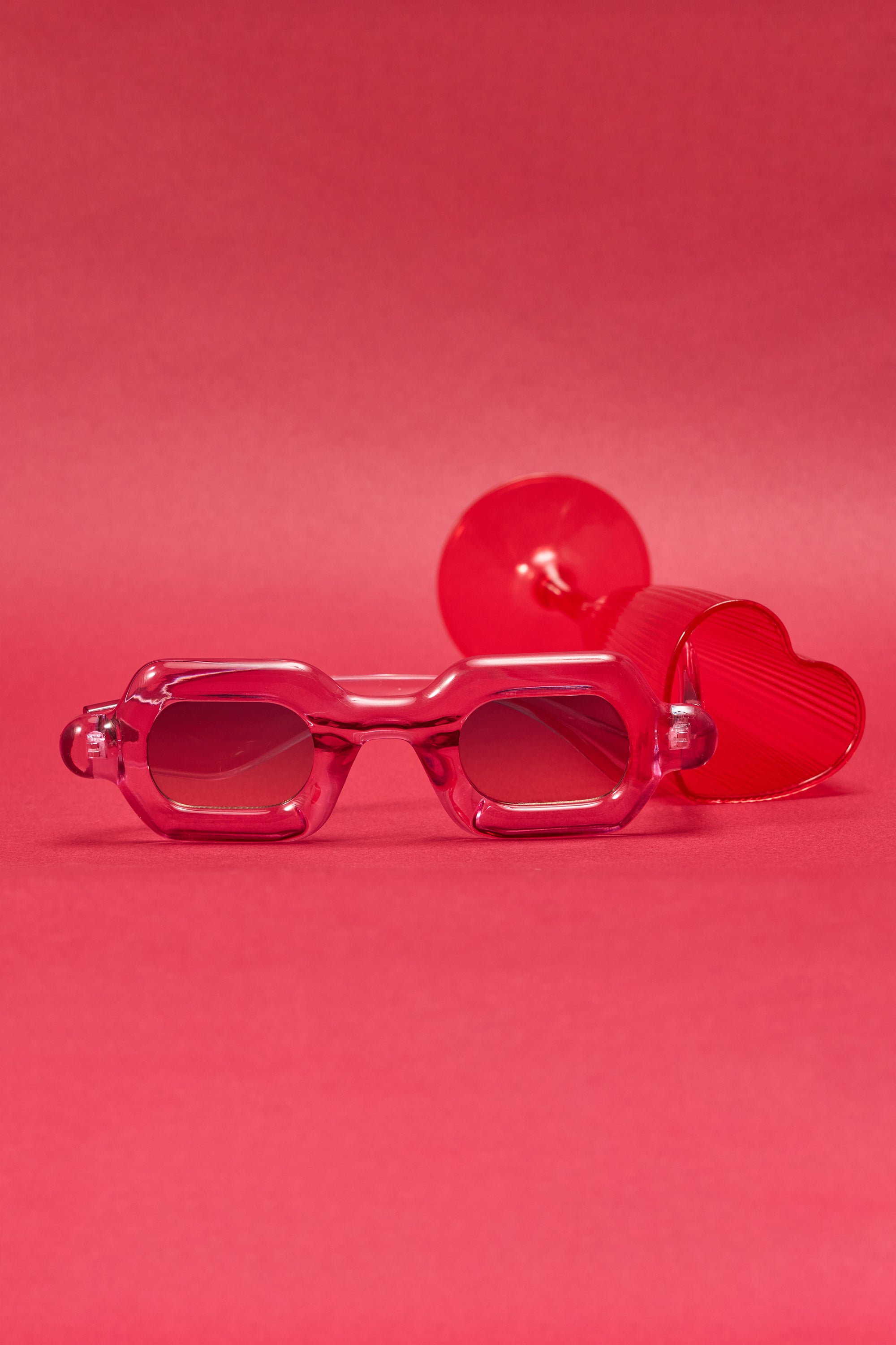 JELLY SUNGLASSES with chunky pink frames and a red heart-shaped accent are displayed on a solid pink background, creating a playful and vibrant scene. 1