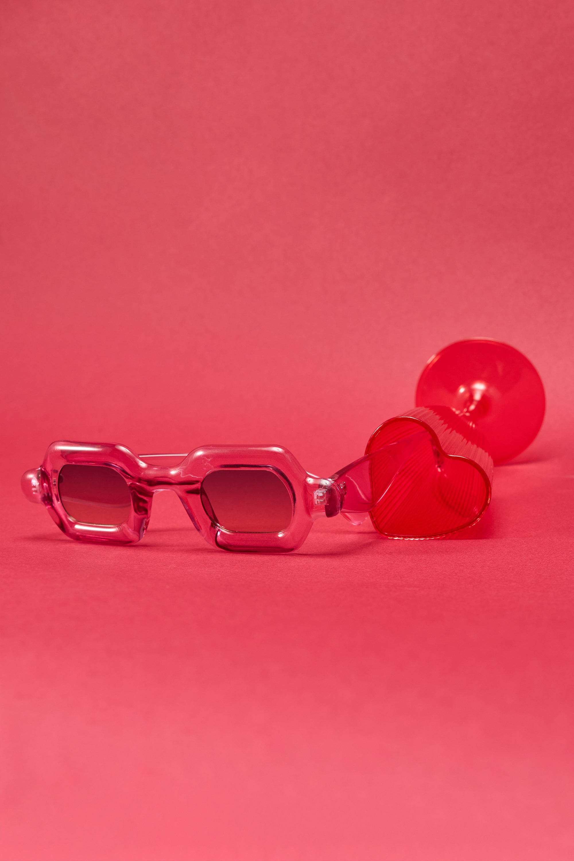 JELLY SUNGLASSES, featuring a chunky pink rectangular frame, rest on a matching pink surface beside a red heart-shaped object, all with a coordinating pink background. 4