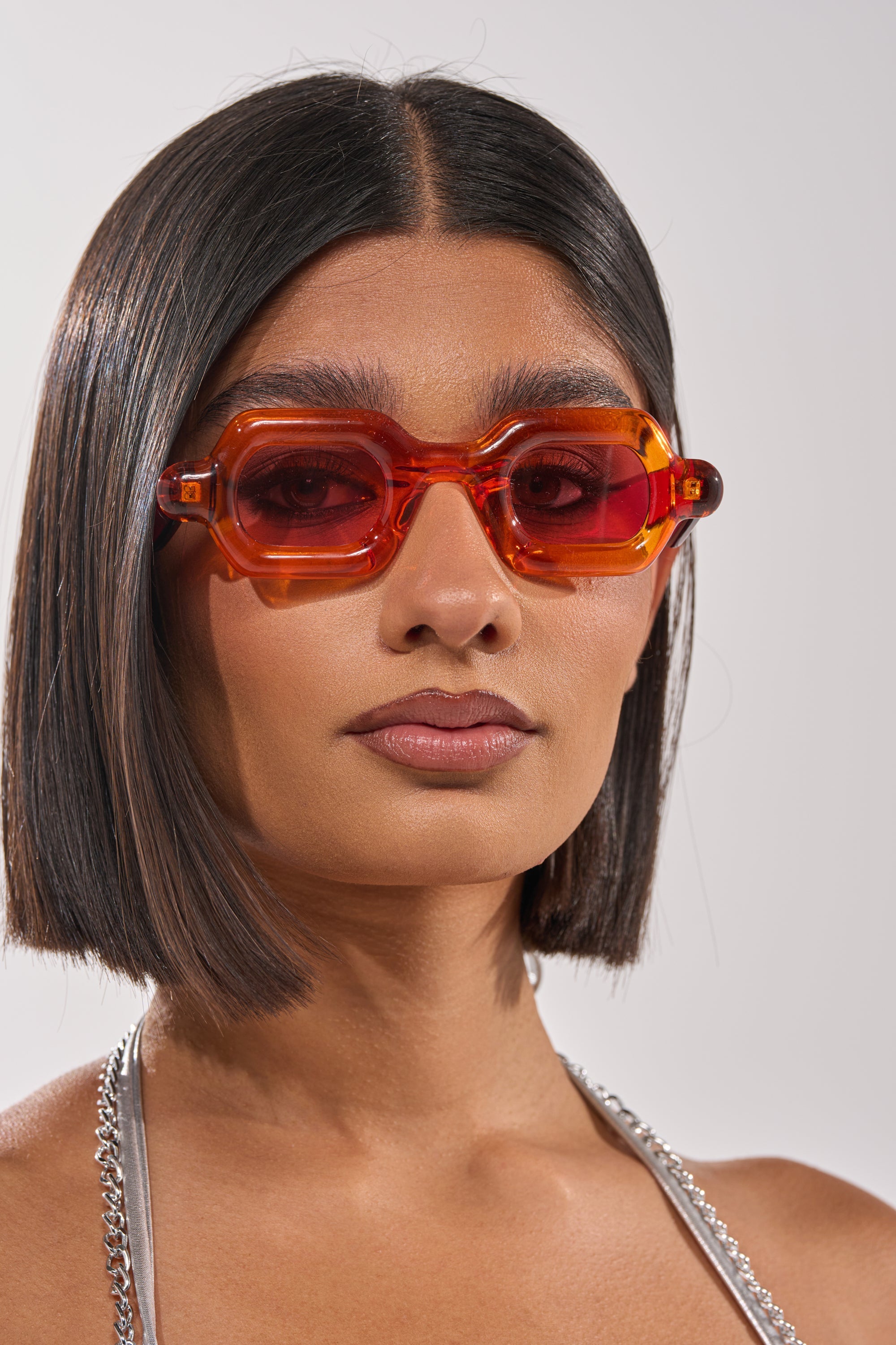 A woman with straight, shoulder-length dark hair wears SORBET SUNGLASSES—chunky orange frames with rosy tinted lenses—and a silver chain-strap top, gazing confidently at the camera against a plain background. 1
