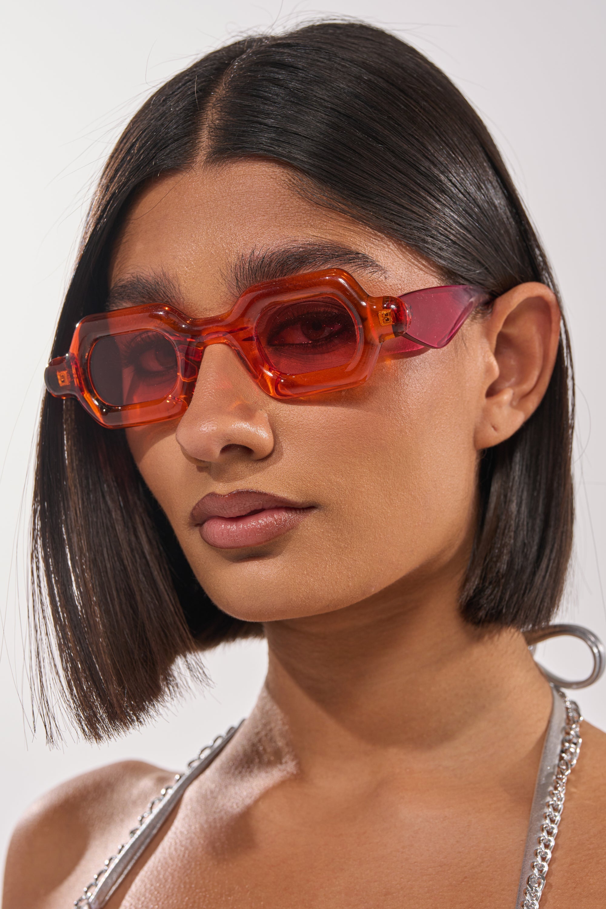 A person with straight, shoulder-length dark hair wears bold, chunky SORBET SUNGLASSES and a metallic halter top, gazing confidently at the camera against a plain background. 2