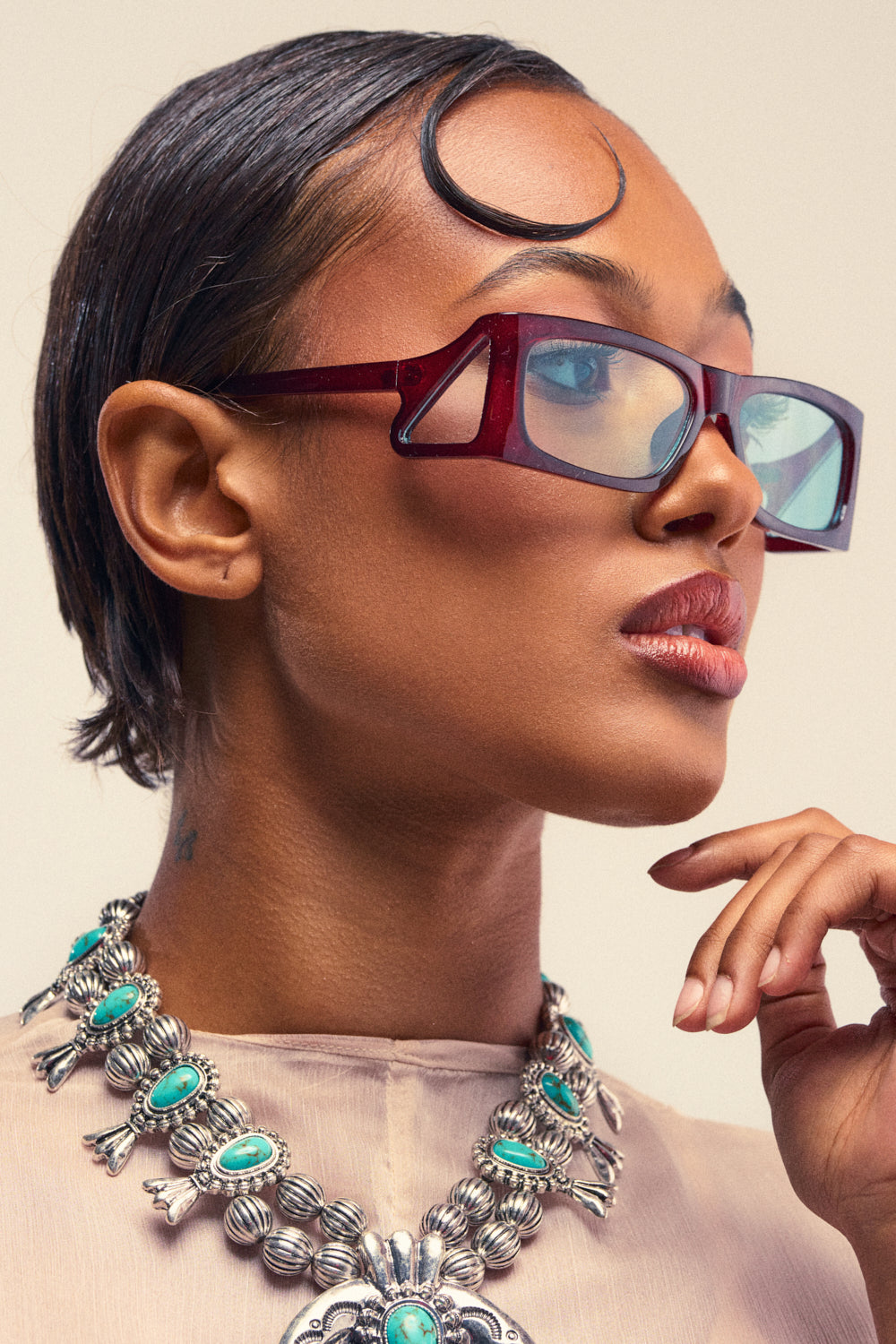 Wearing the KEKE SUNGLASSES, a woman with slicked-back hair and a sheer top accessorizes her bold look with a large silver necklace featuring turquoise stones as she gazes to the side.