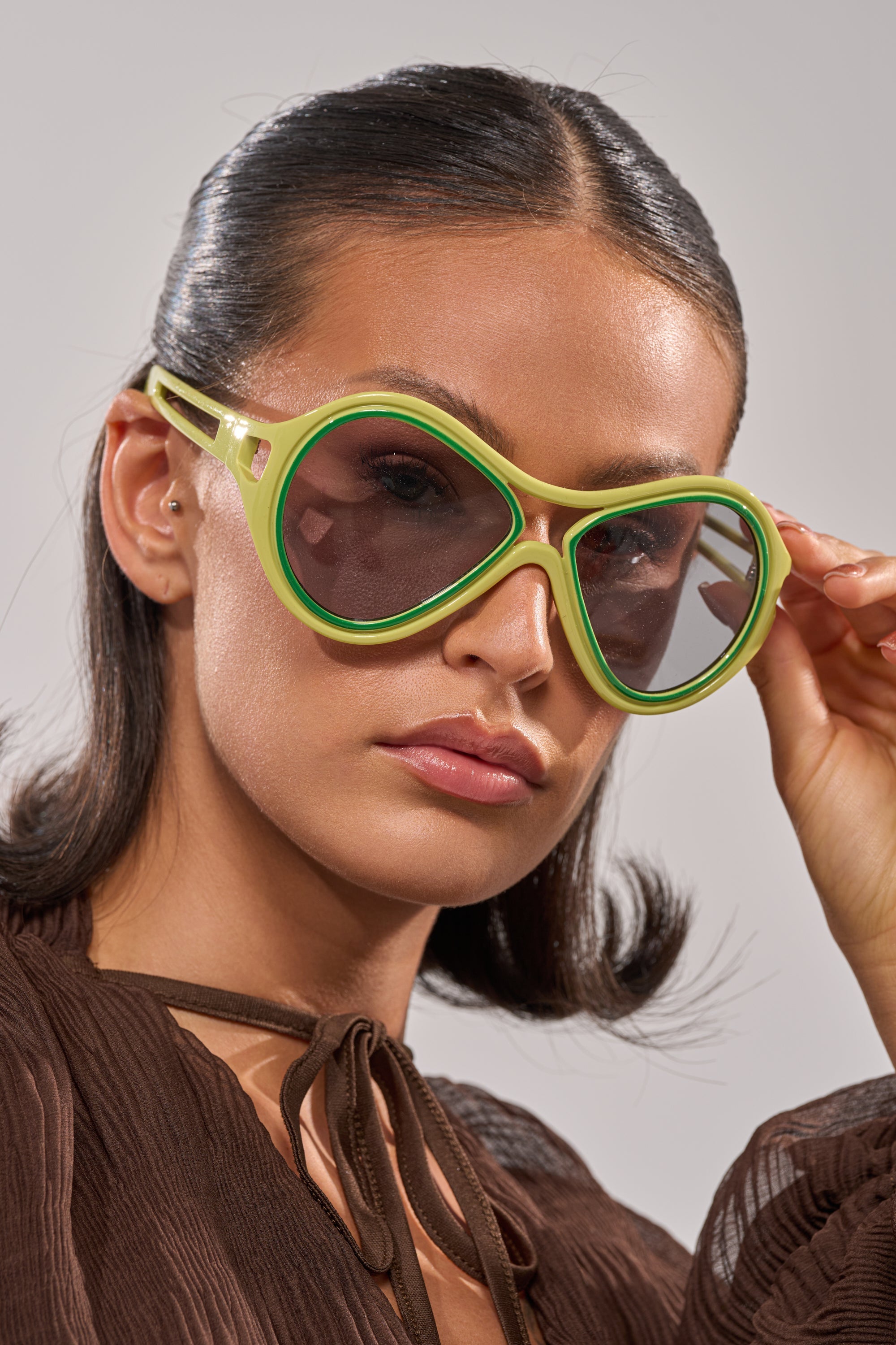 A woman with smooth dark hair wears the IN MY WORLD SUNGLASSES—large, retro green ovals—and a brown textured top, looking confidently at the camera as she holds one side of her glasses with her right hand. 1