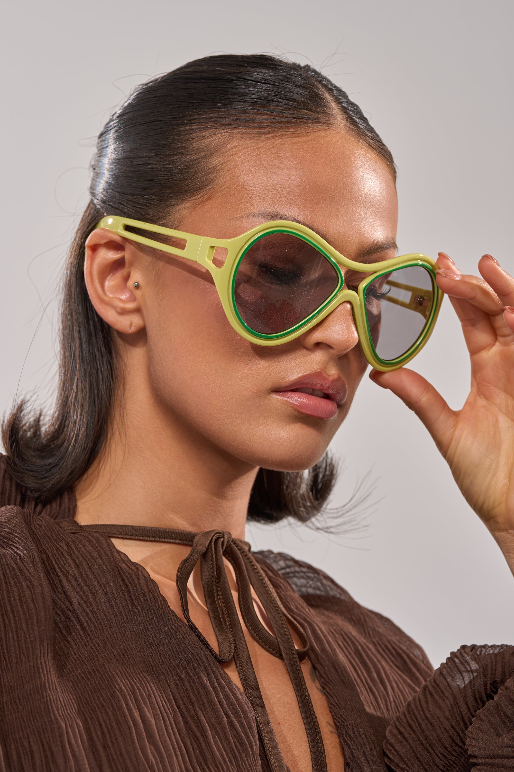 A woman with sleek dark hair stylishly adjusts her IN MY WORLD SUNGLASSES—oversized, retro-inspired with green rims—while wearing a brown, textured long-sleeve blouse tied at the neck and gazing to the side. 2