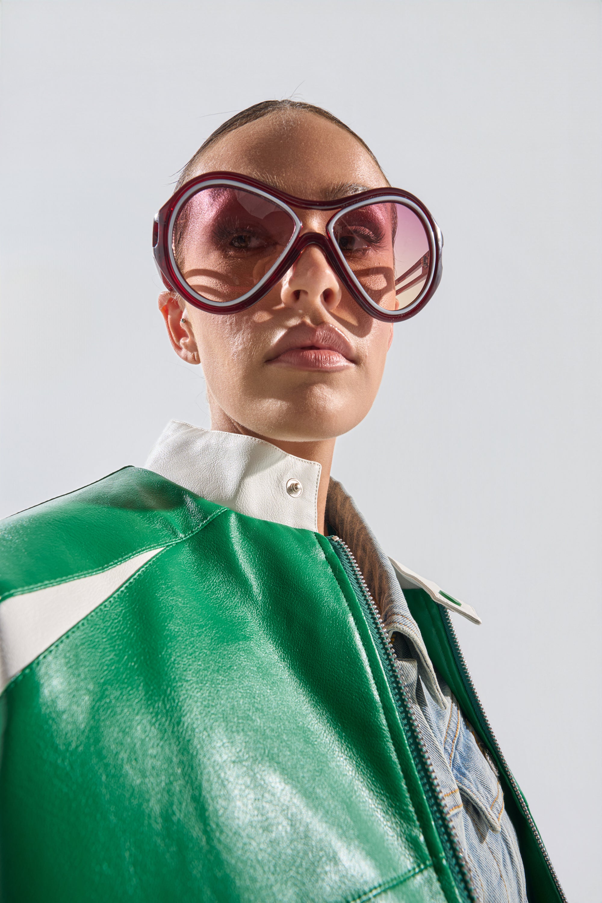 A person wearing WILLY SUNGLASSES with oversized, pink-tinted lenses, paired with a green and white leather jacket and denim shirt, confidently looks at the camera against a plain light background. 1