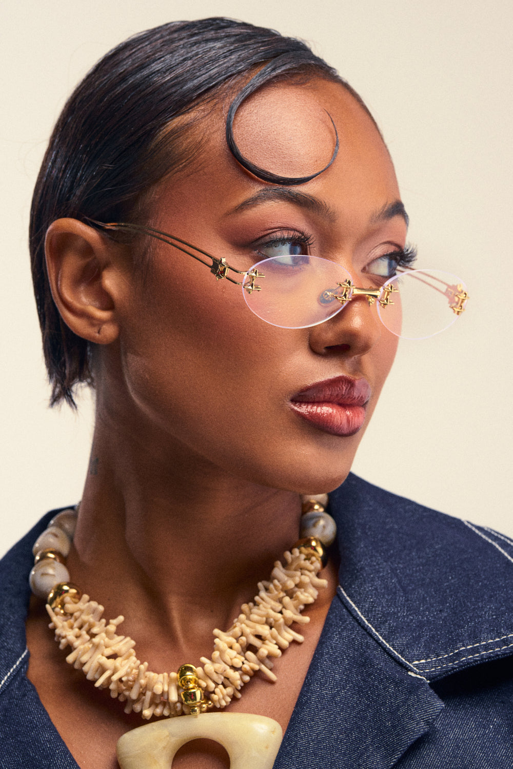 A woman with slicked-back hair and a curled strand wears the minimalist rimless U SEE ME GLASSES with clear lenses, paired with a chunky beige and gold necklace and a dark denim jacket, looking off to the side.