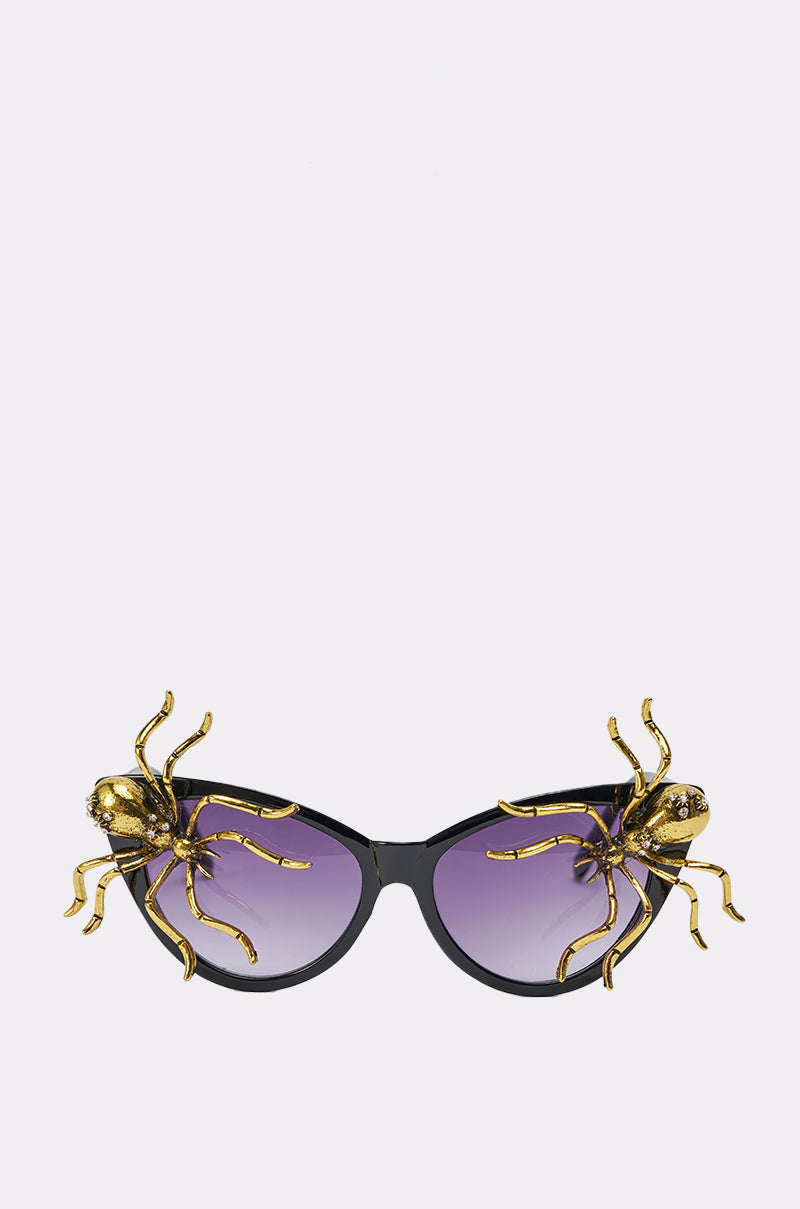 BITE ME SUNGLASSES are black cat-eye frames with purple lenses, accented by large gold spider sculptures on each top corner—a bold statement accessory shown against a plain white background. 0