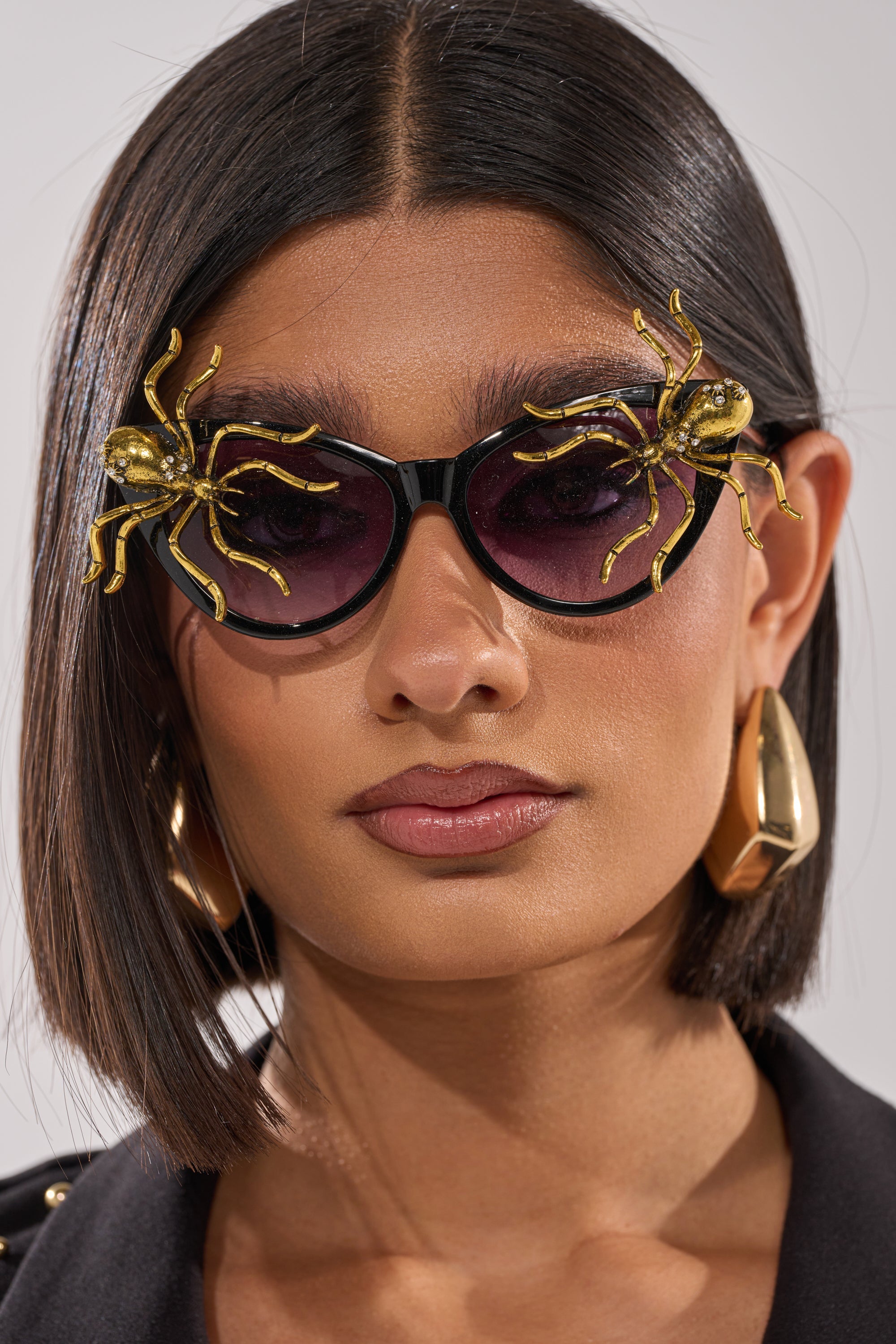 A woman with straight, dark hair wears large gold earrings and chic BITE ME SUNGLASSES accented with gold spider embellishments. Dressed in black, she gazes at the camera, her look completed by bold Halloween-inspired accessories. 1