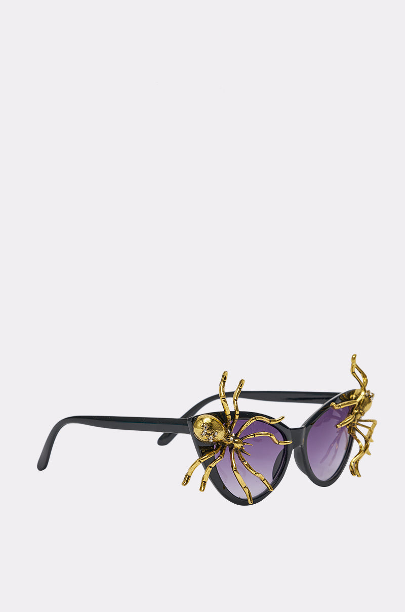 The BITE ME SUNGLASSES feature black cat-eye frames, purple lenses, and bold gold spider accents on the rims, creating a dramatic statement against a simple light background. 4