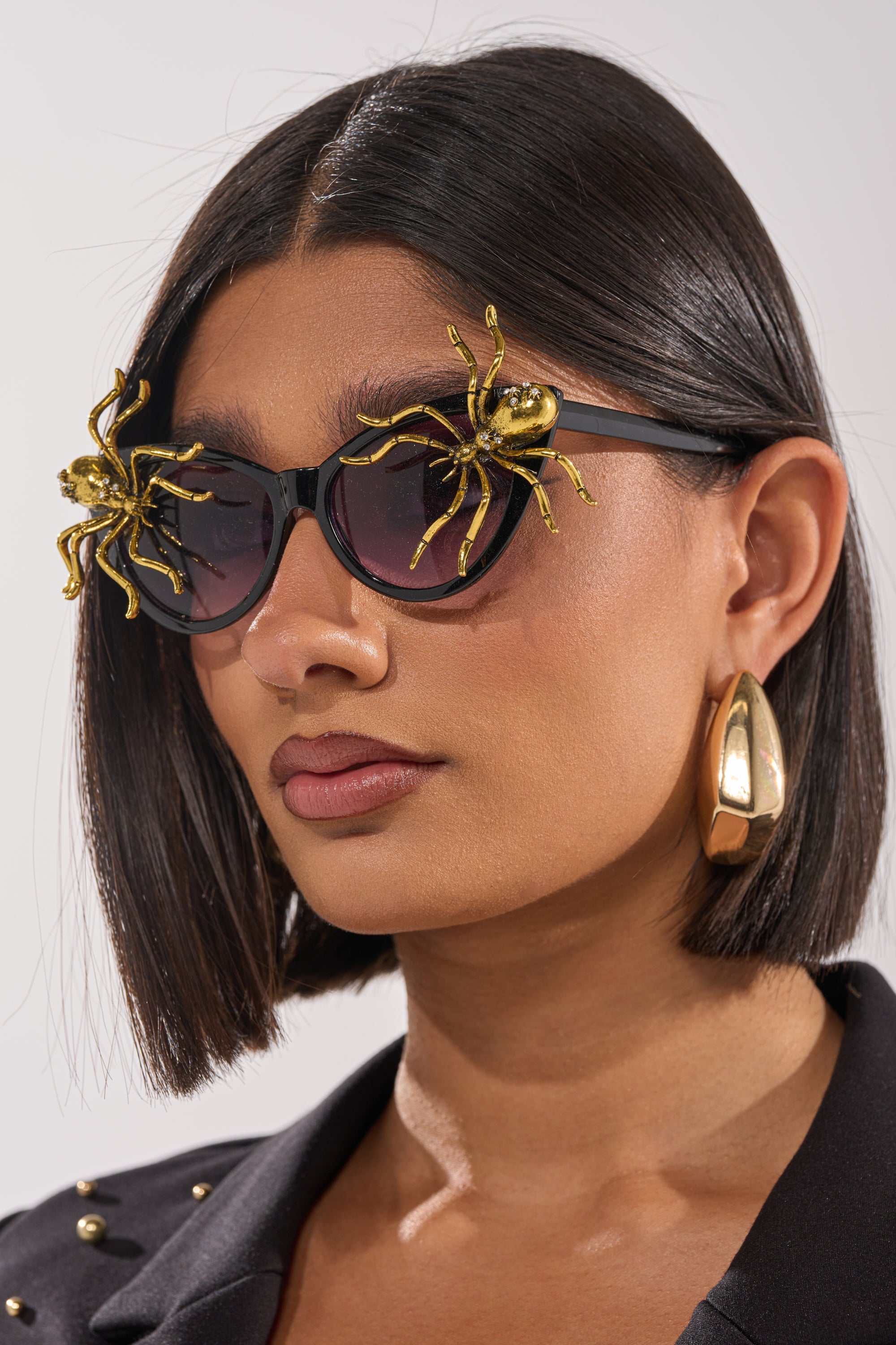 A woman with straight, dark bobbed hair wears BITE ME SUNGLASSES, black cat-eye frames adorned with large gold spider embellishments. She also sports chunky gold hoop earrings and a black studded blazer. 2
