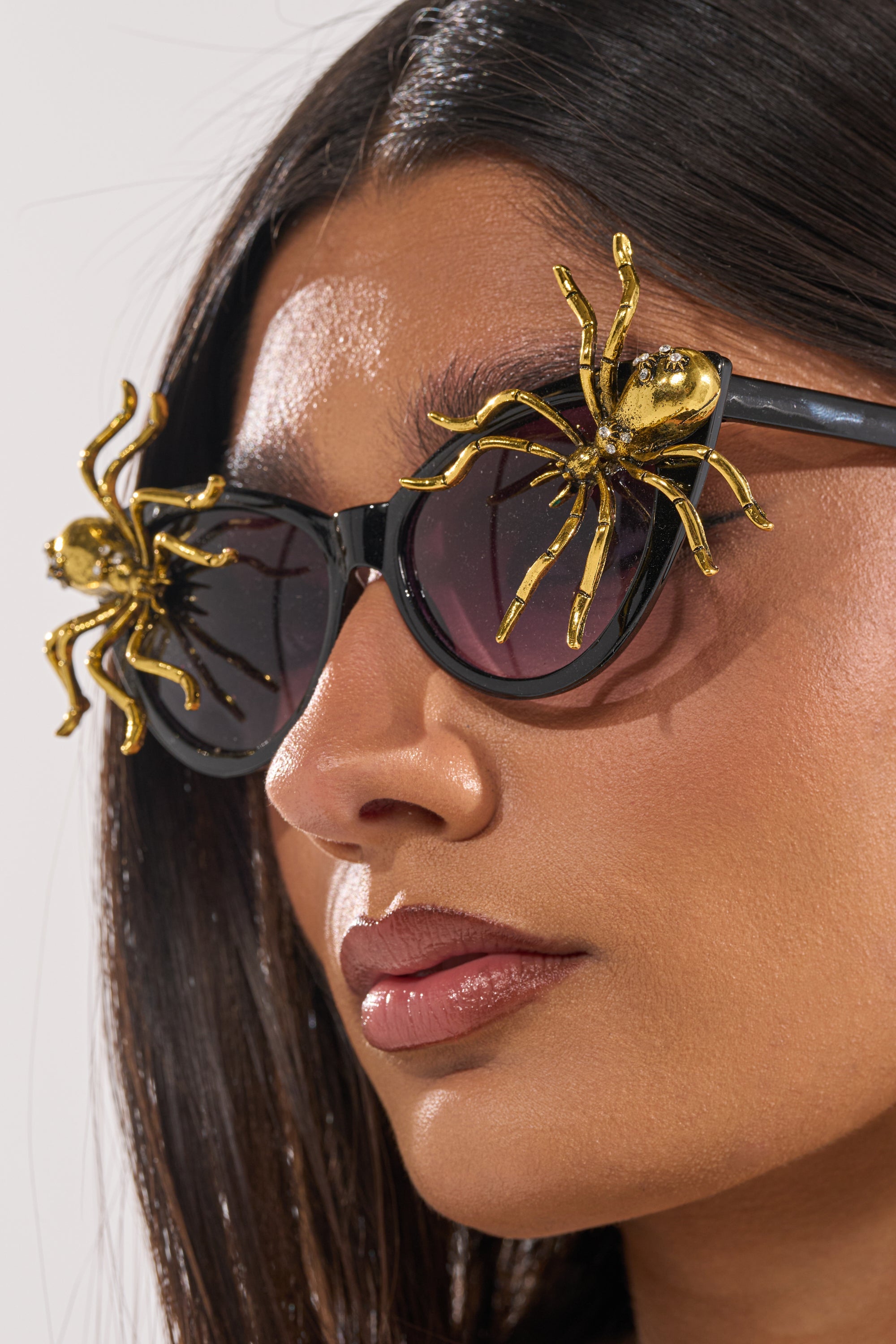 A woman with smooth dark hair and a neutral expression wears BITE ME SUNGLASSES—black cat-eye frames topped with large gold spider accents on each lens—making these bold Halloween accessories truly stand out. 3