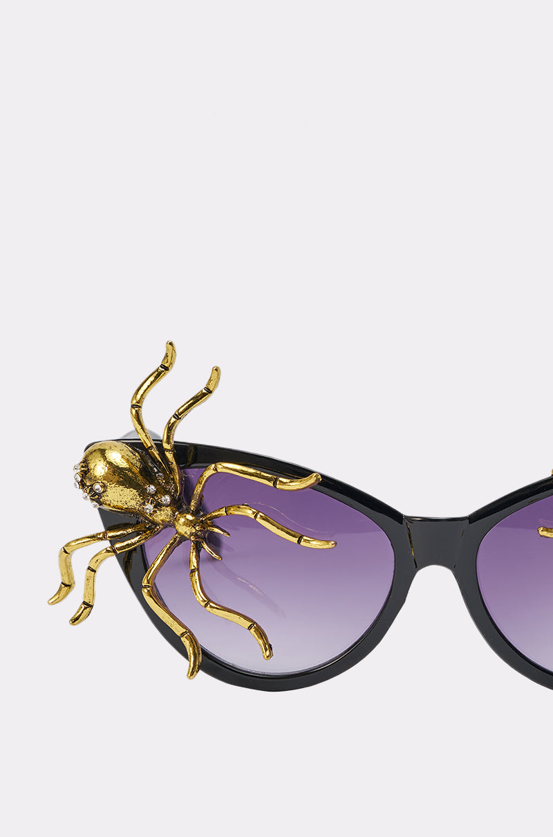 The BITE ME SUNGLASSES are bold black cat-eye frames with purple lenses, adorned with a large jeweled gold spider on one corner for a striking statement look. 5