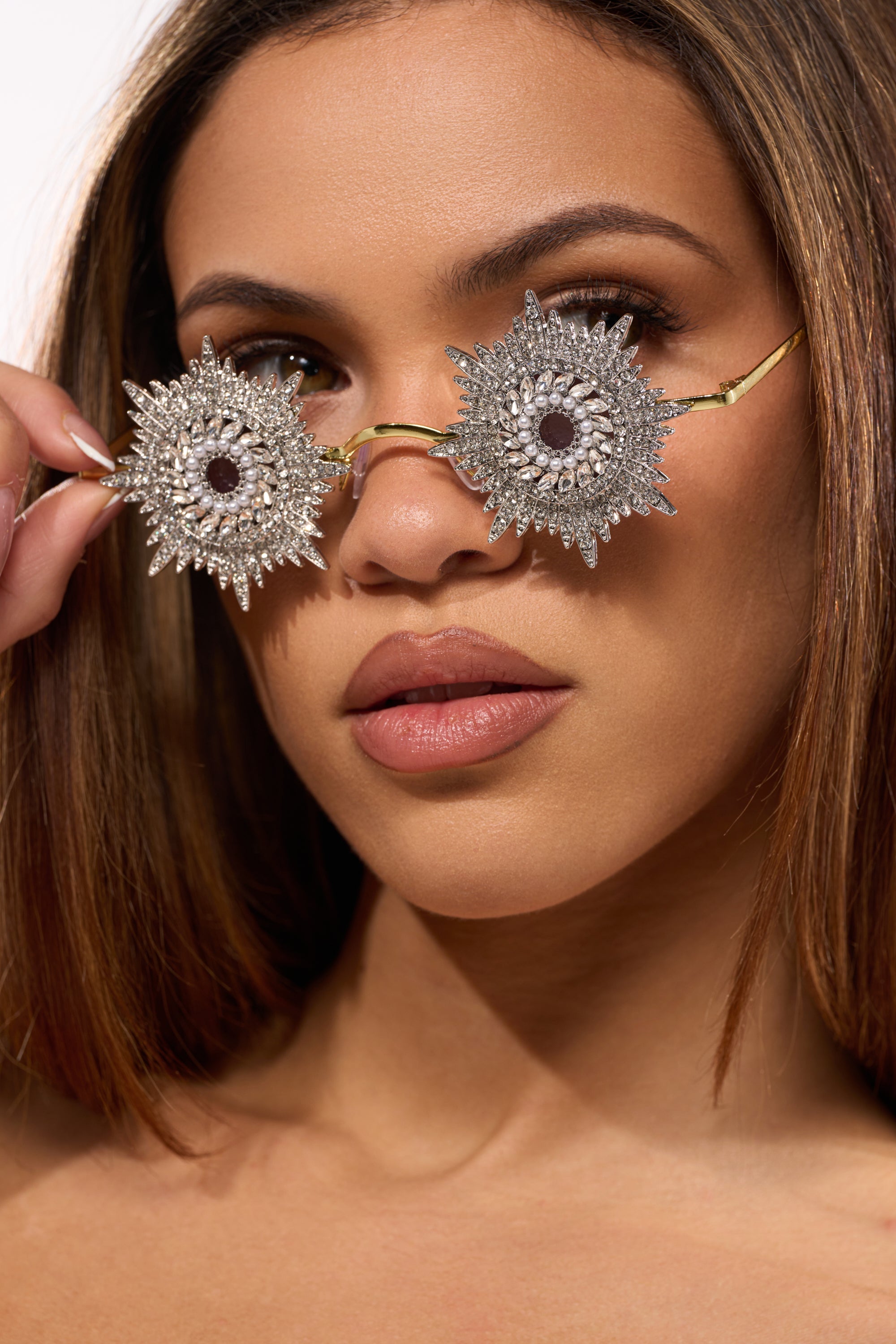 A woman with straight brown hair lifts her WHY DO I NEED TO SEE SUNGLASSES, which feature large, circular, silver sunburst-shaped frames with intricate details, revealing her brown eyes and neutral-toned makeup. 1