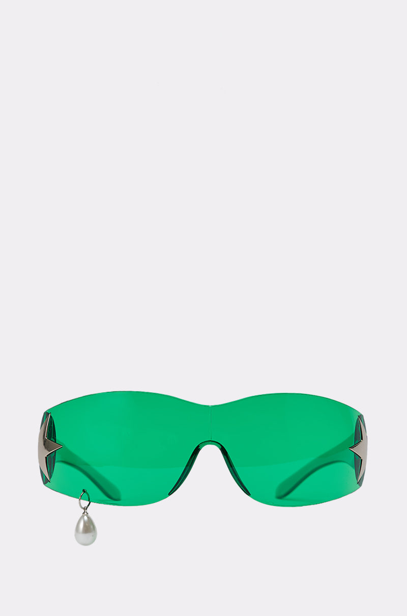 The I DONT CRY SUNGLASSES feature green wraparound shield lenses, silver star-shaped accents on each side, and a dangling pearl charm on the left, giving them a futuristic edge against a light-colored background. 0