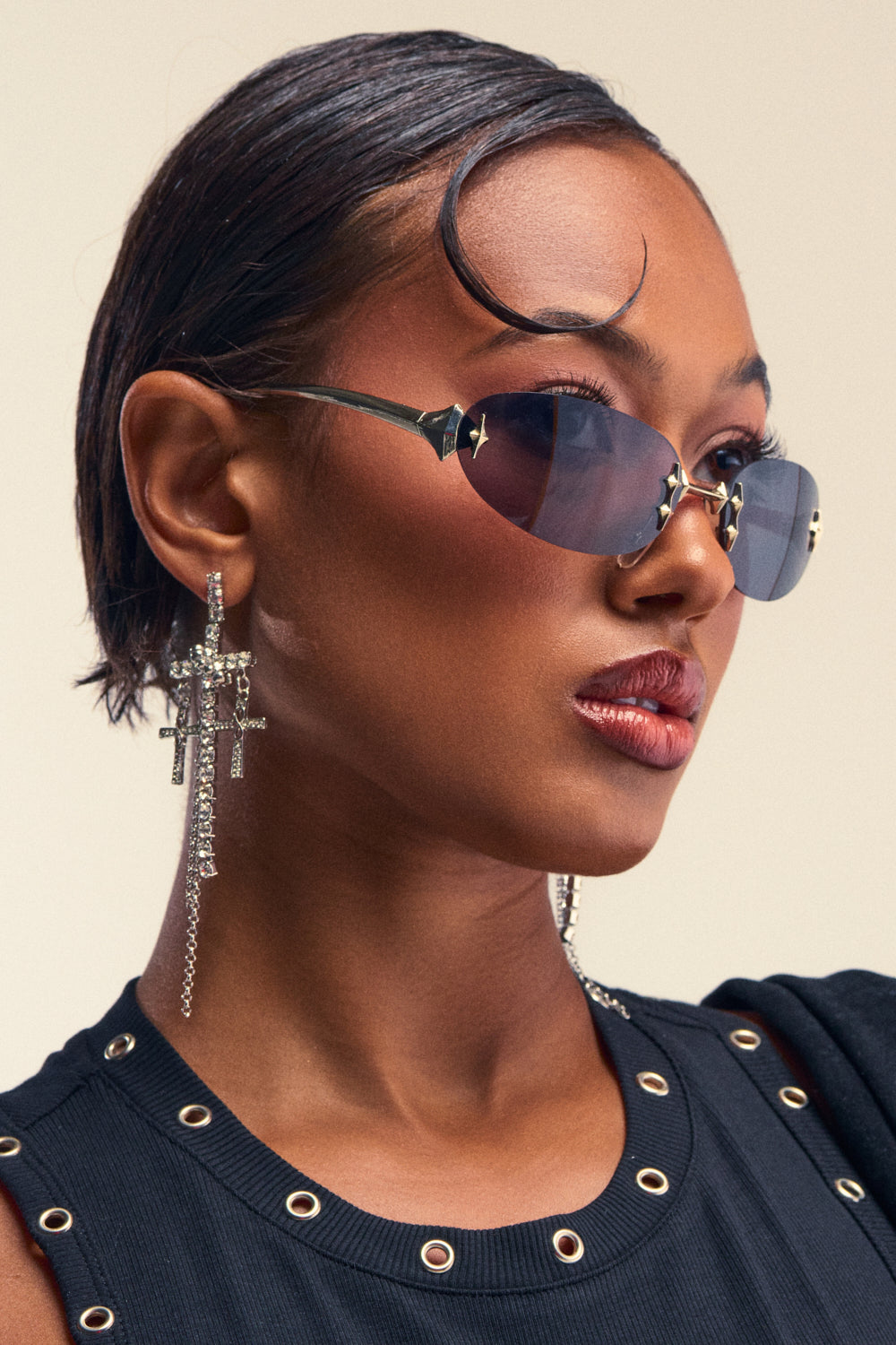 A close-up profile shot of a woman with slicked-back dark hair, wearing CYBER SUNGLASSES—Y2K-inspired rimless oval blue-tinted sunglasses—paired with long dangling crystal cross earrings and a black top with silver grommets around the neckline, posing against a neutral background.