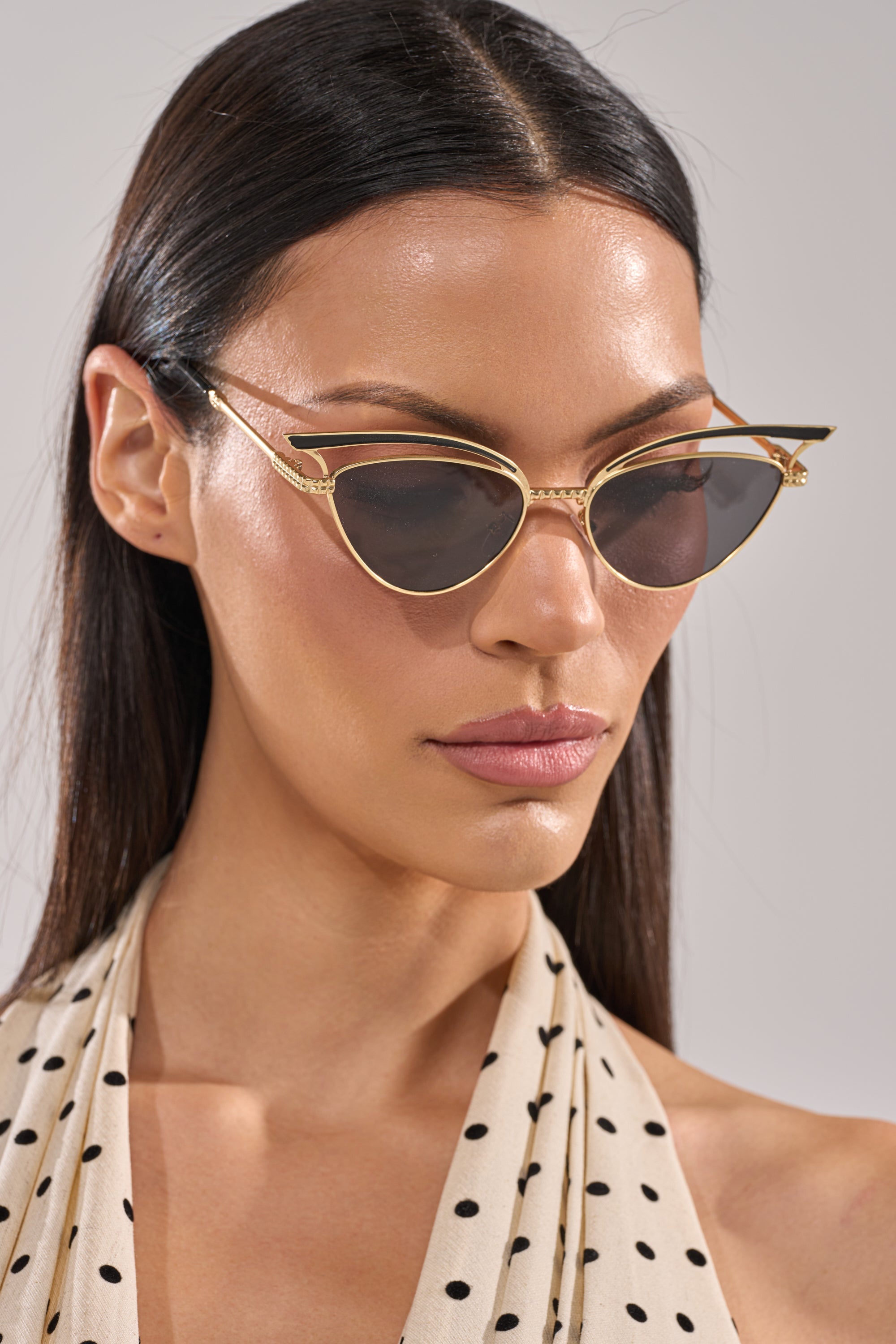 A woman with straight dark hair wears AUDREY SUNGLASSES and a cream halter top with black polka dots, looking downward with a neutral expression against a plain background. 1