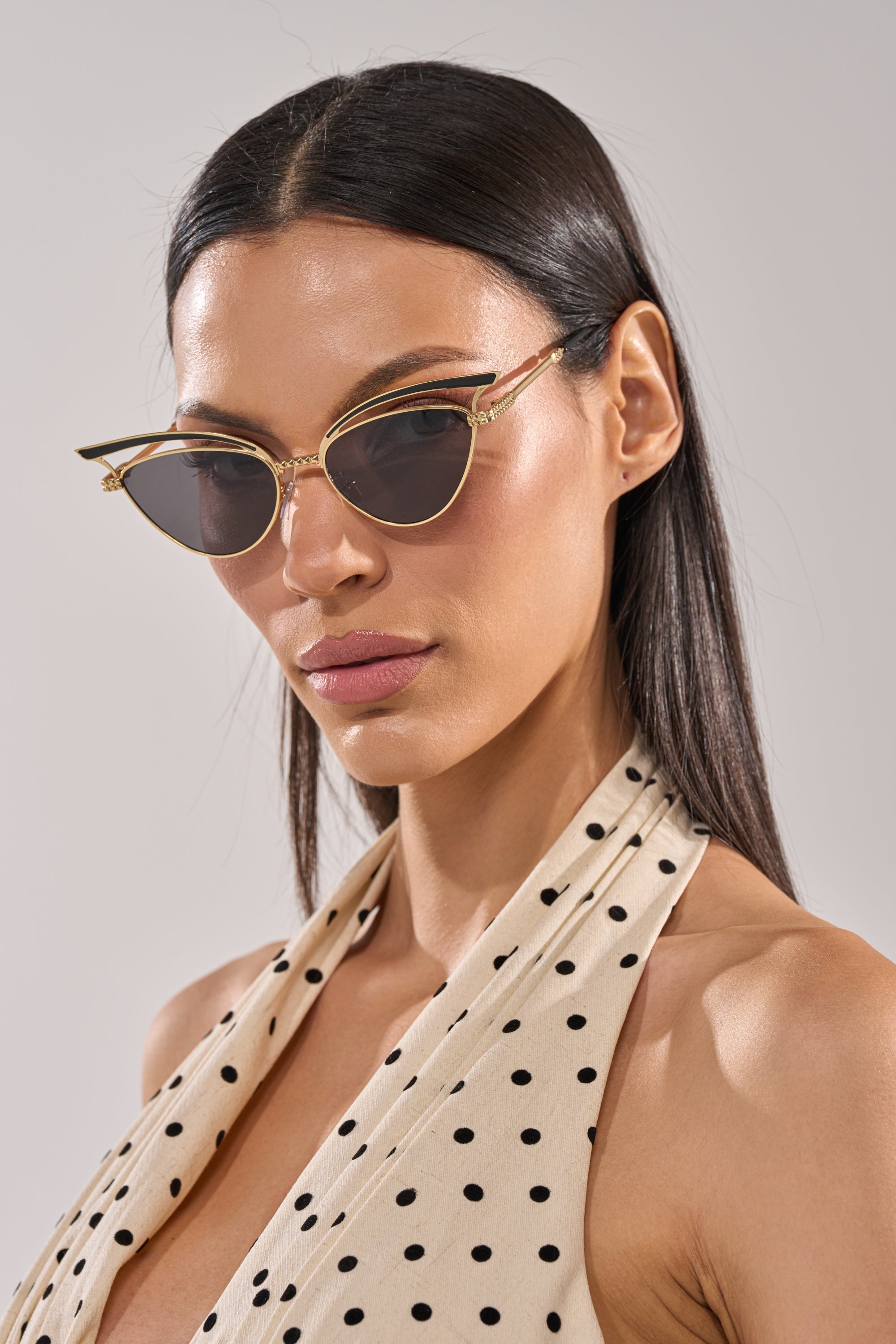 A woman with straight dark hair wears the AUDREY SUNGLASSES, featuring gold-accented cat-eye frames and dark lenses, with a cream halter top with black polka dots, posing against a light gray background. 2